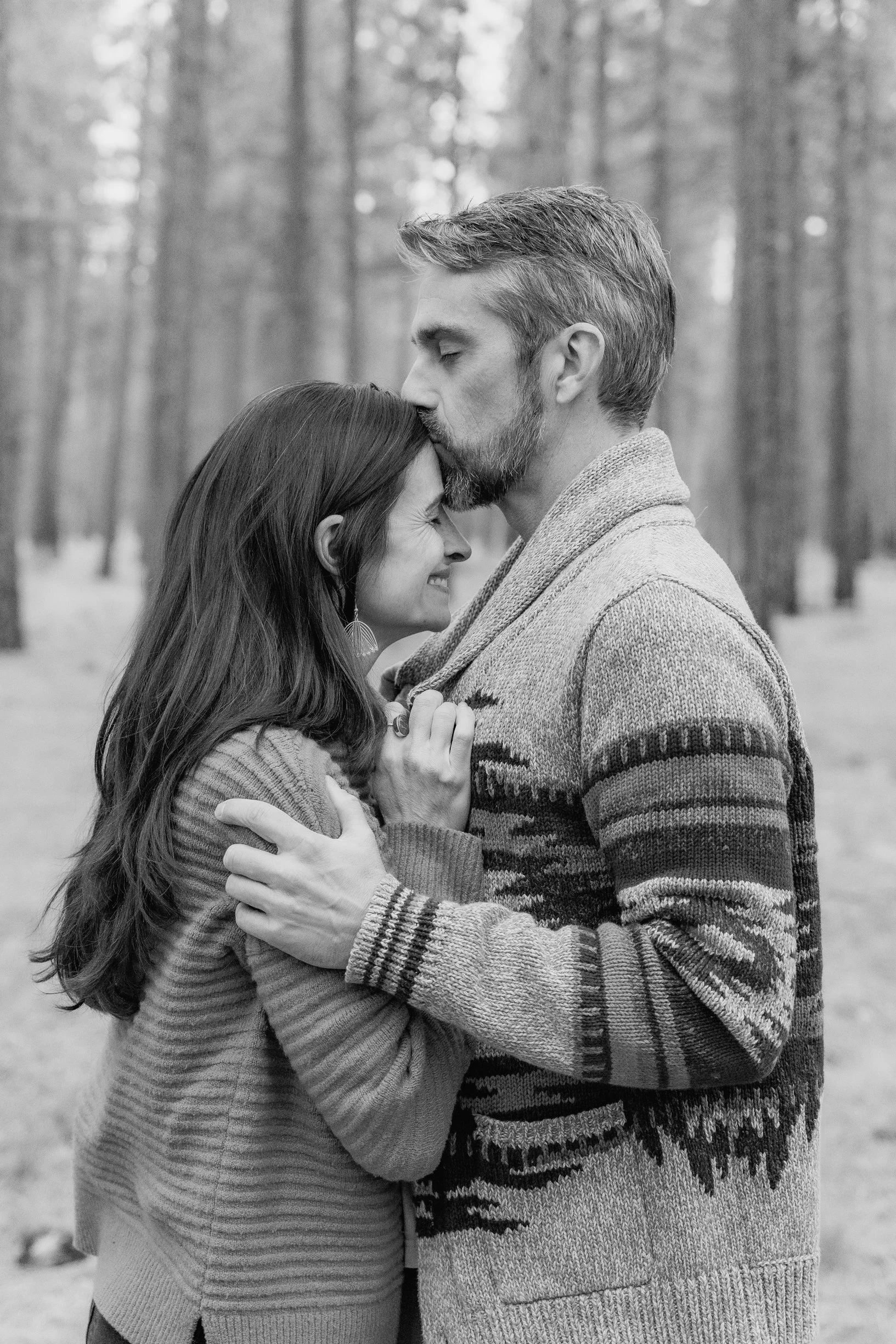 A black and white photo of a couple embracing and sharing a kiss in a forest. The woman is smiling with her eyes closed, and the man is gently kissing her forehead. They are wearing sweaters, and the background shows tall trees.