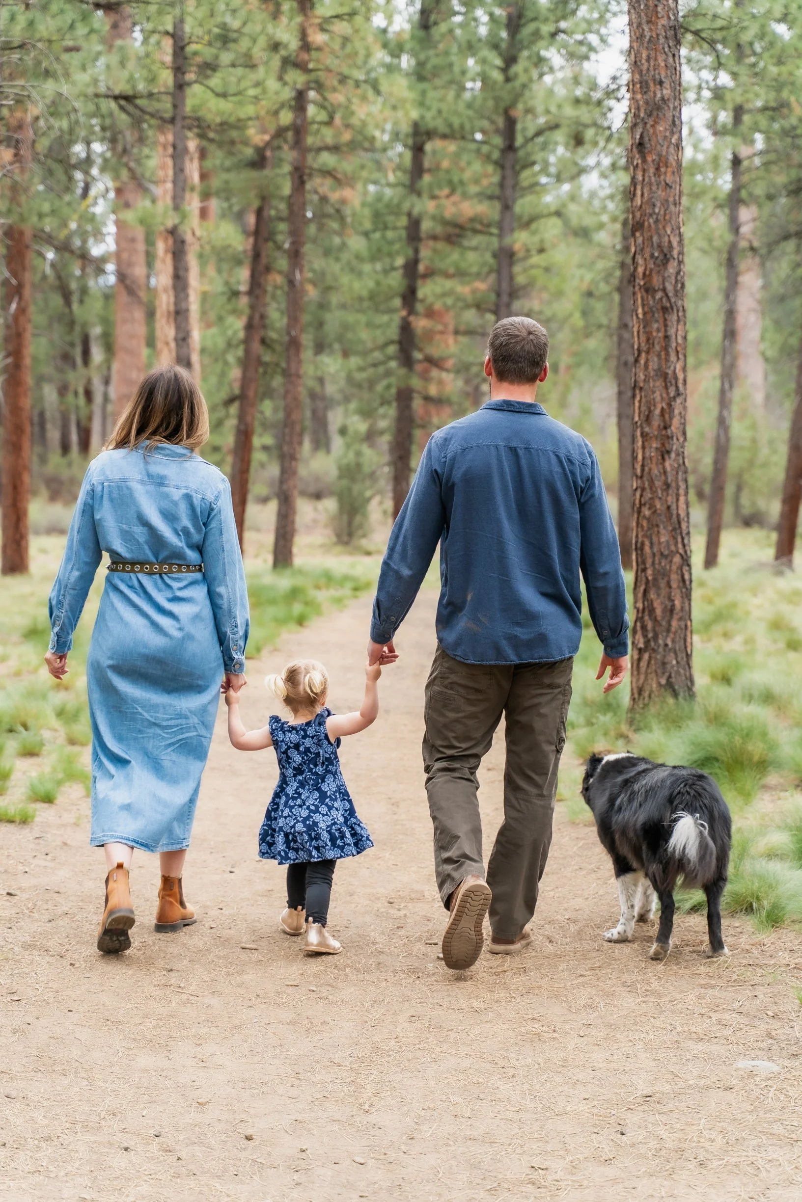 man and woman walk down a path in the forest with their daughter between then and their dog at their side.