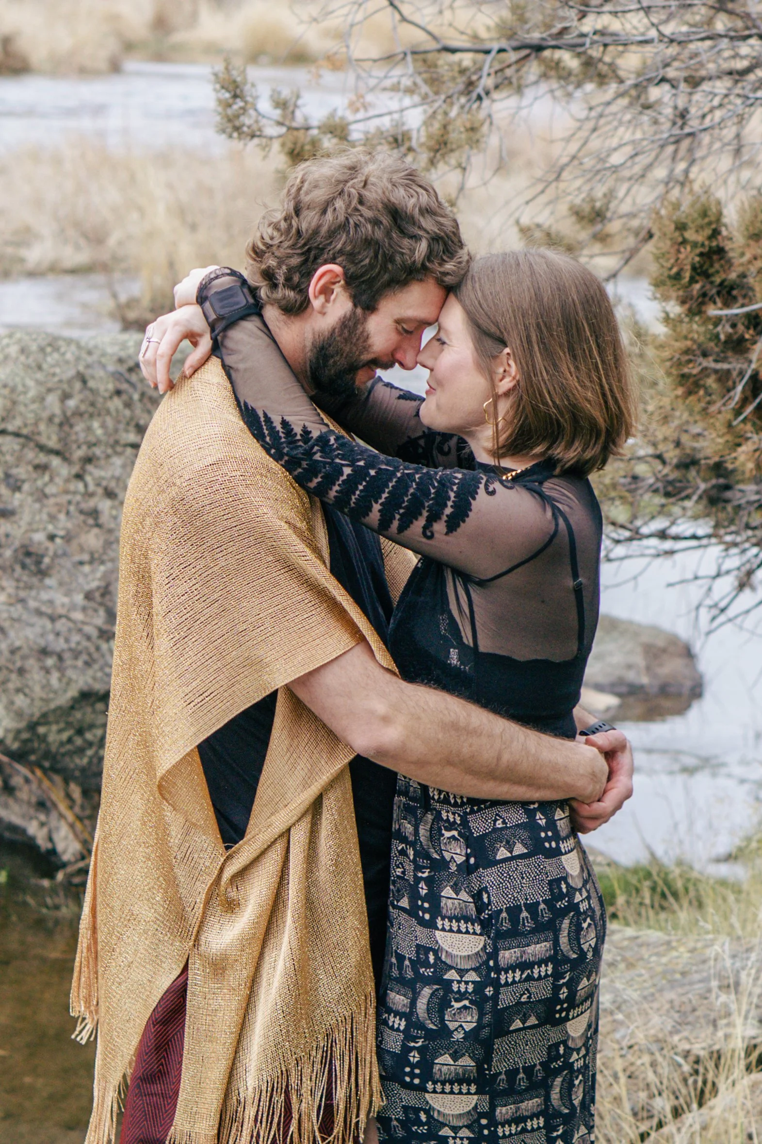 A man and woman embrace with foreheads touching outdoors by a river, surrounded by rocks and dry vegetation.
