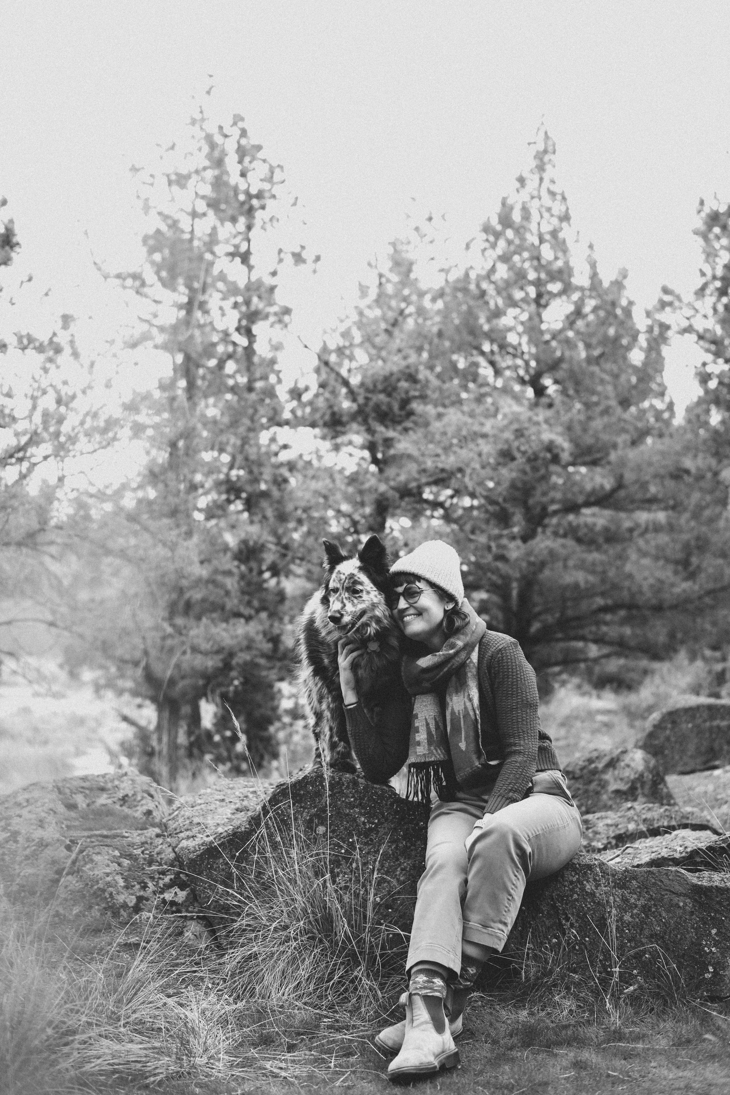 A woman sitting on a rock in a forest, holding a dog with a multi-colored coat, surrounded by tall trees and grass.