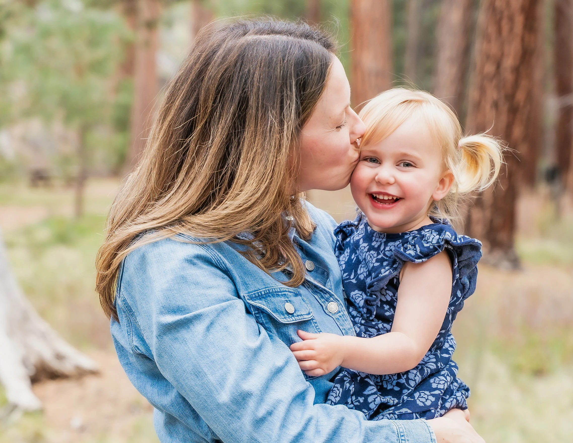 A woman with long brown hair holding a smiling blonde girl with pigtails, outdoors in a wooded area.