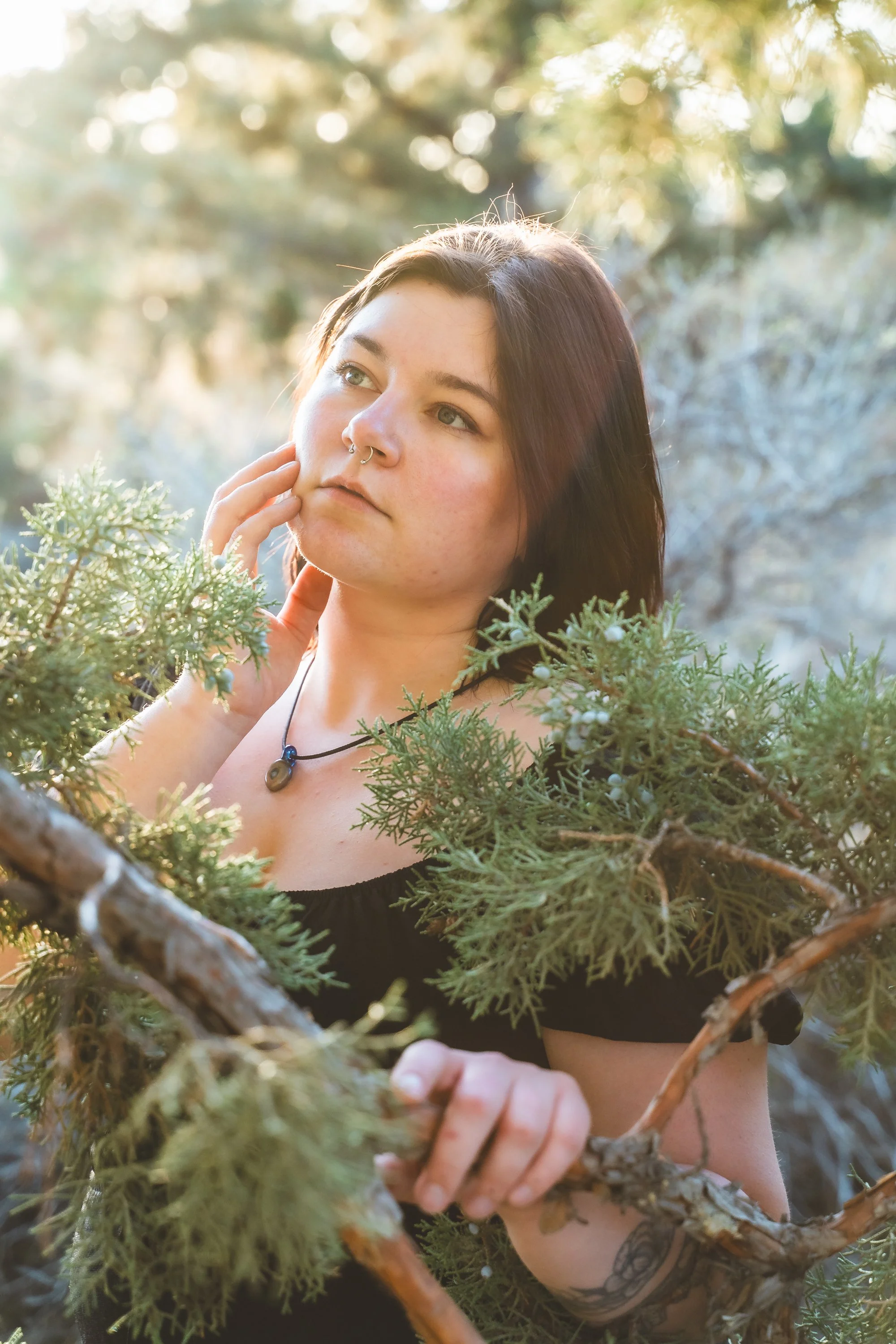 A woman with dark hair, a septum piercing, and tattoos, sitting outdoors surrounded by green trees and branches, gazing thoughtfully into the distance.