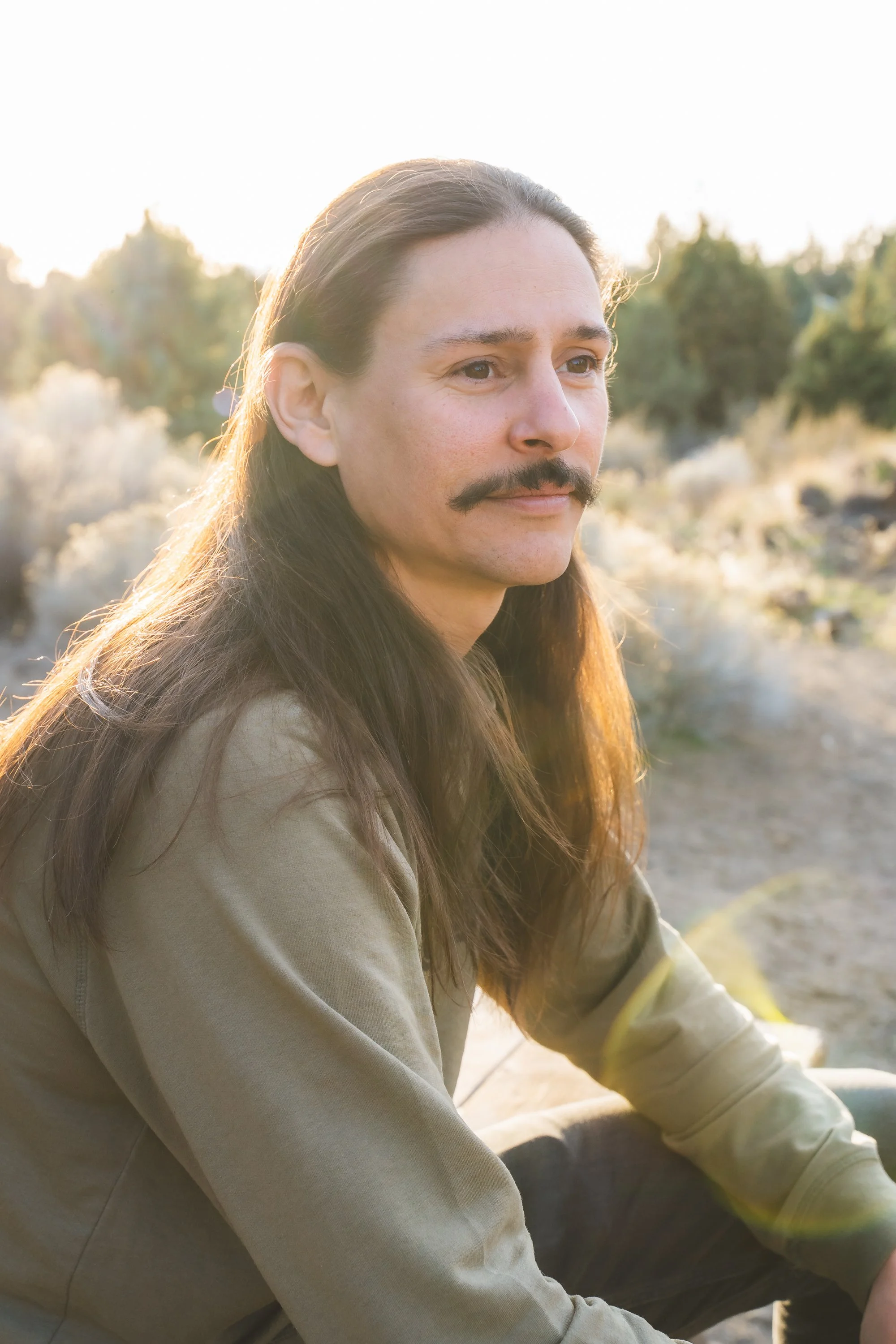 A man with long brown hair, a mustache, and light skin sitting outdoors in sunlight, wearing a light-colored jacket, with trees in the background.
