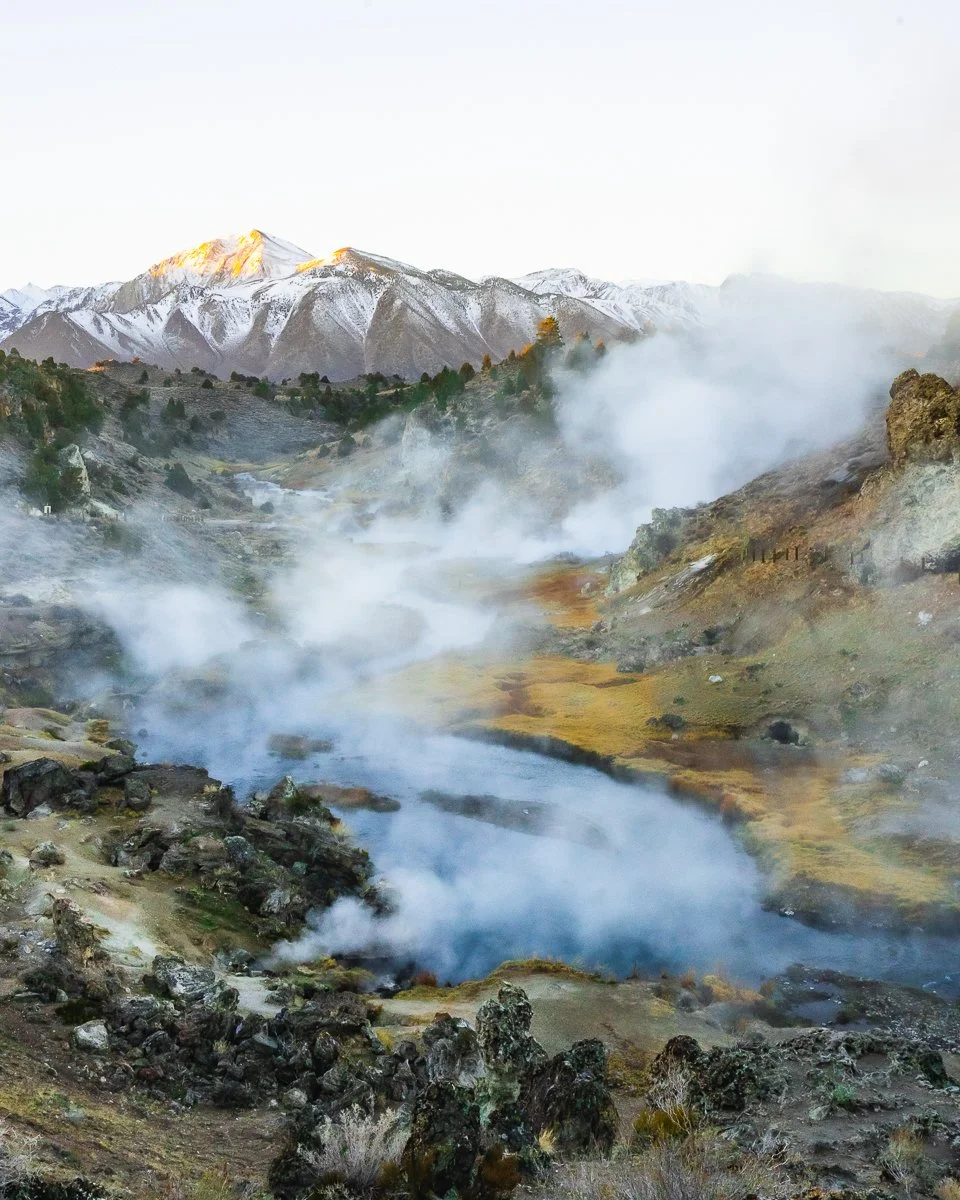 Mountain landscape with snow-capped peaks, steam rising from geothermal vents, and a rugged terrain.
