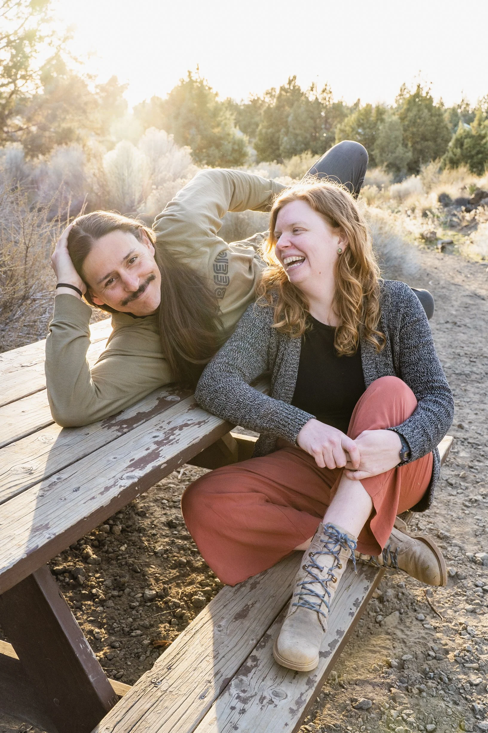 Two friends, a man with a mustache and long hair, and a woman with curly red hair, laughing and relaxing on a wooden picnic table outdoors during sunset, surrounded by trees and shrubs.