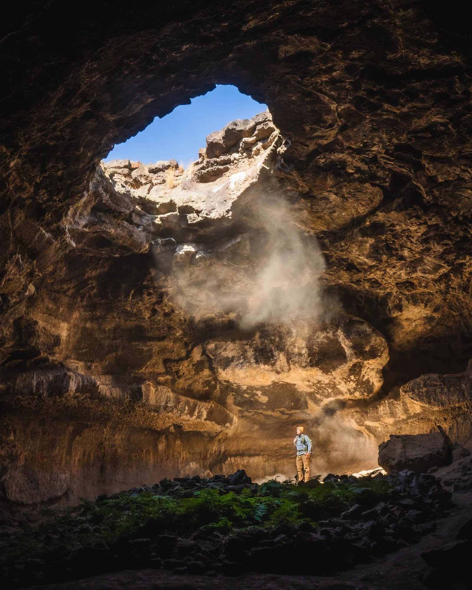 A person with a backpack and hat standing inside a large natural cave or cavern with a rocky ceiling. Sunlight streams in through a hole in the ceiling, illuminating the person and the green plants on the ground near them. The scene suggests explorat