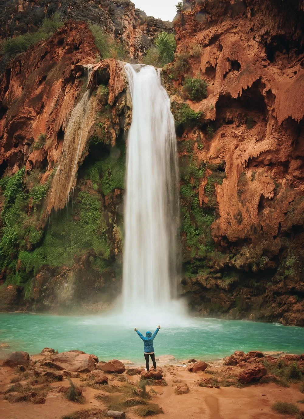 A person with arms raised standing on rocks by a turquoise pool at the base of a large waterfall cascading down red rocky cliffs surrounded by greenery.