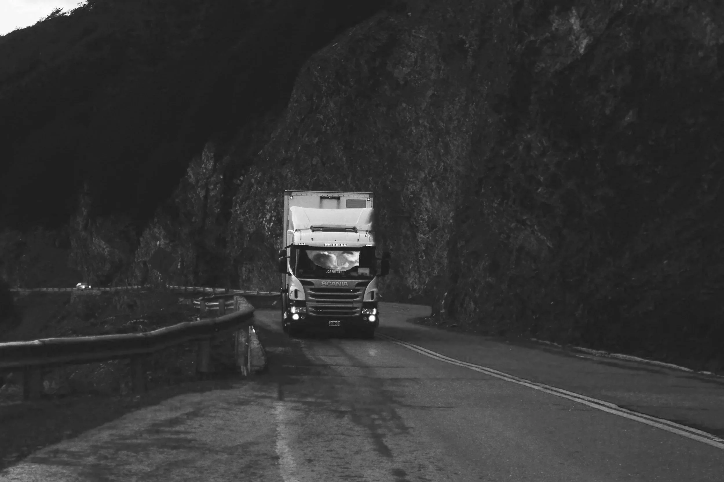 A white truck driving on a winding mountain highway with rocky cliffs on one side and a guardrail on the other, under an overcast sky.