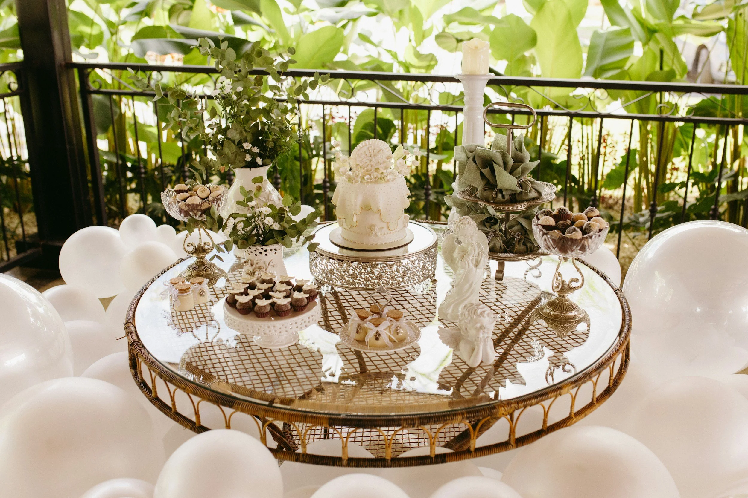 Table decorated for a celebration with a white cake, chocolate cupcakes, and various sweets, surrounded by white balloons, greenery, and a balcony in the background.