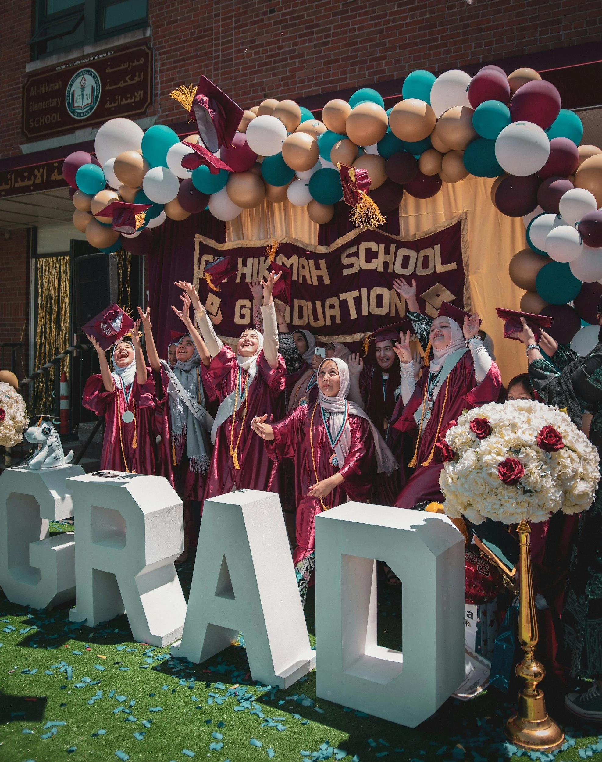 A graduation celebration at Al-Hikmah Elementary School with students in maroon caps and gowns tossing caps in the air, decorated with balloons and flowers, on a stage with a banner that reads 'HIKMAH SCHOOL GRADUATION'.