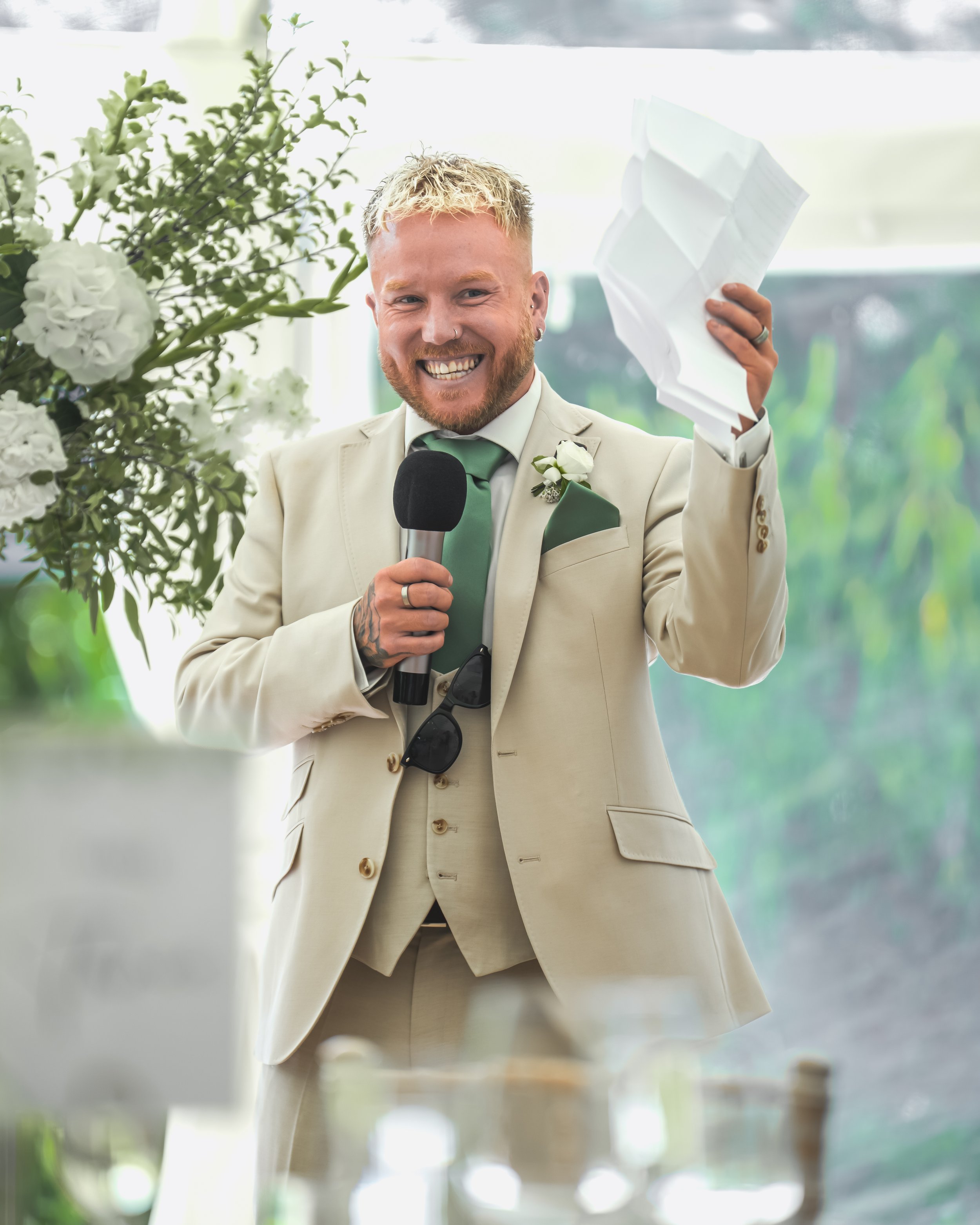 A smiling man in a light beige suit holding a microphone and a crumpled piece of paper, standing in front of a large window with greenery outside, decorated with white flowers.