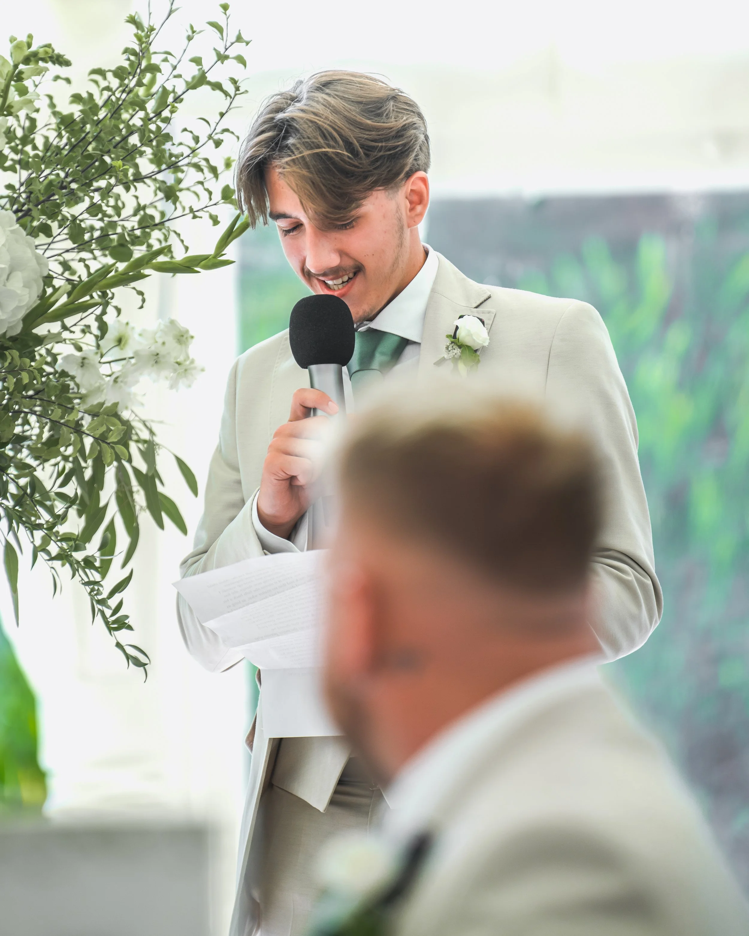 Young man in a light-colored suit giving a speech at an event, holding a microphone, with floral decorations and greenery nearby.