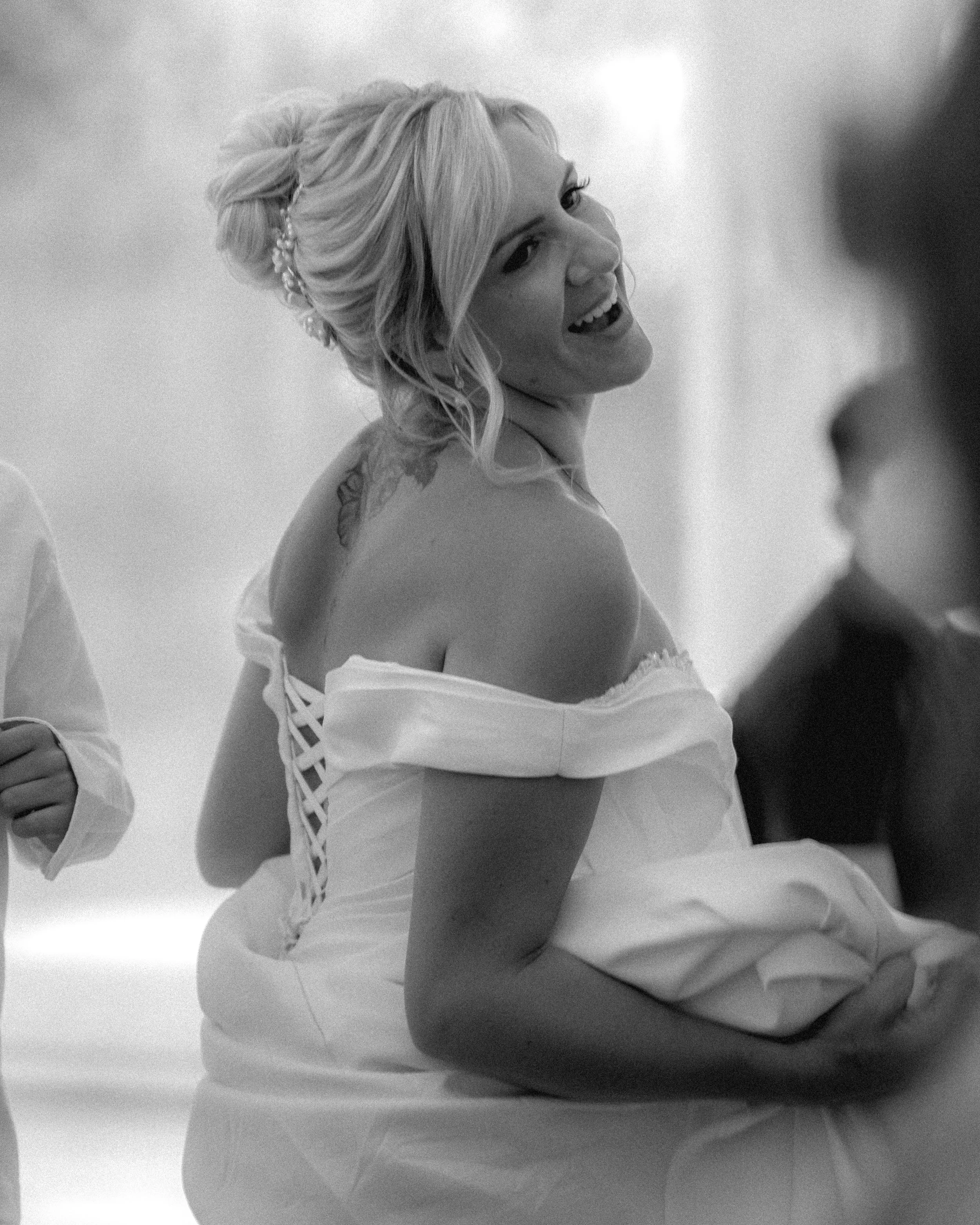 A woman with blonde hair styled in an elegant updo with a pearl hair accessory. She is smiling and looking to the right, wearing a strapless dress with a leathe leaf tattoo on her shoulder. The background is blurred.