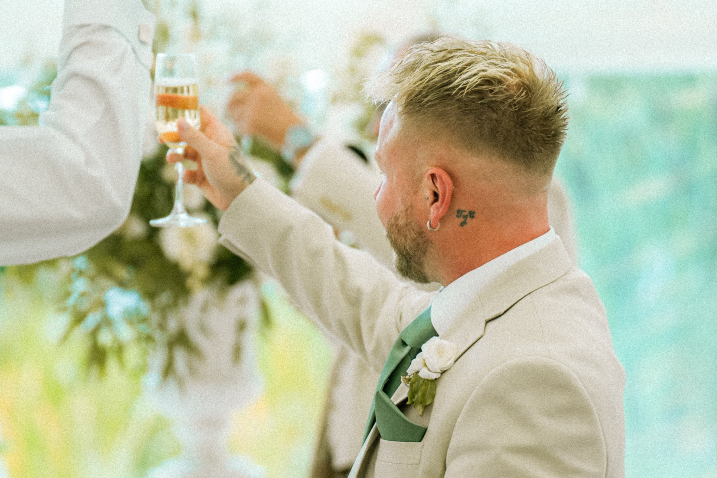 A man with blonde hair and tattoos on his neck, dressed in a beige suit with a pink tie, holding a champagne glass at a formal event.