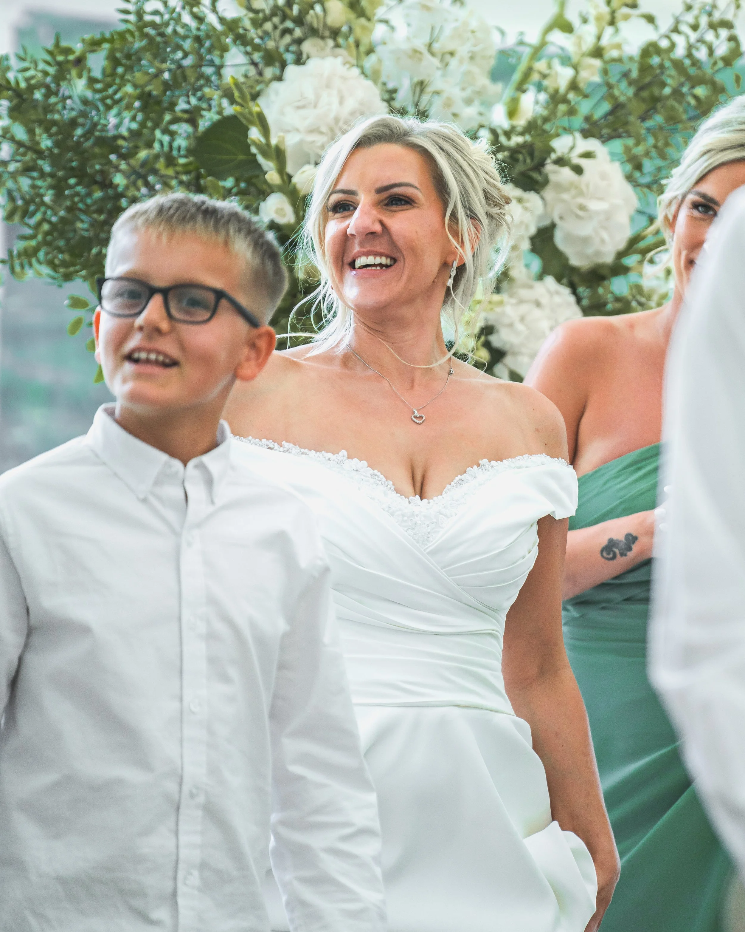 A woman in a white dress smiling at a wedding celebration with a man in glasses and a white shirt next to her, behind a large bouquet of white flowers and greenery.