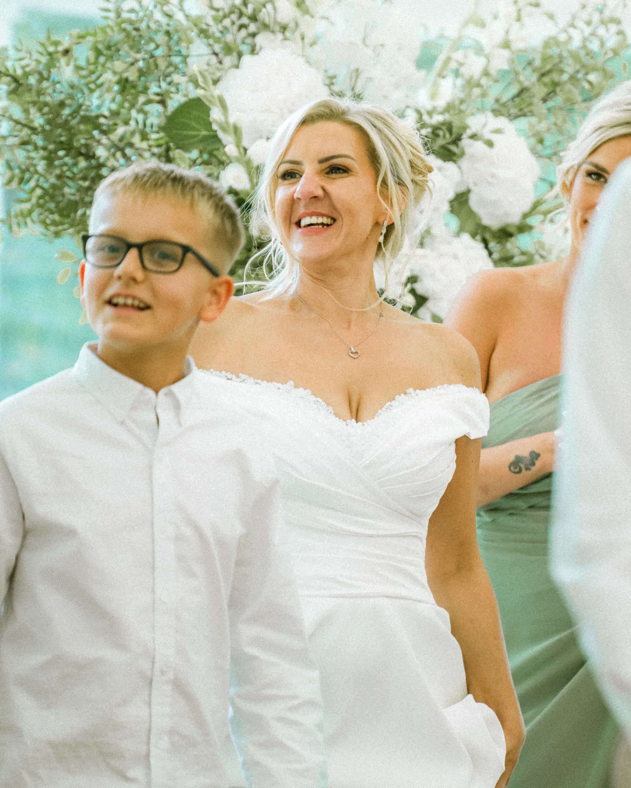 A bride smiling during a wedding ceremony, standing next to groom and another woman, with floral white decorations in the background.