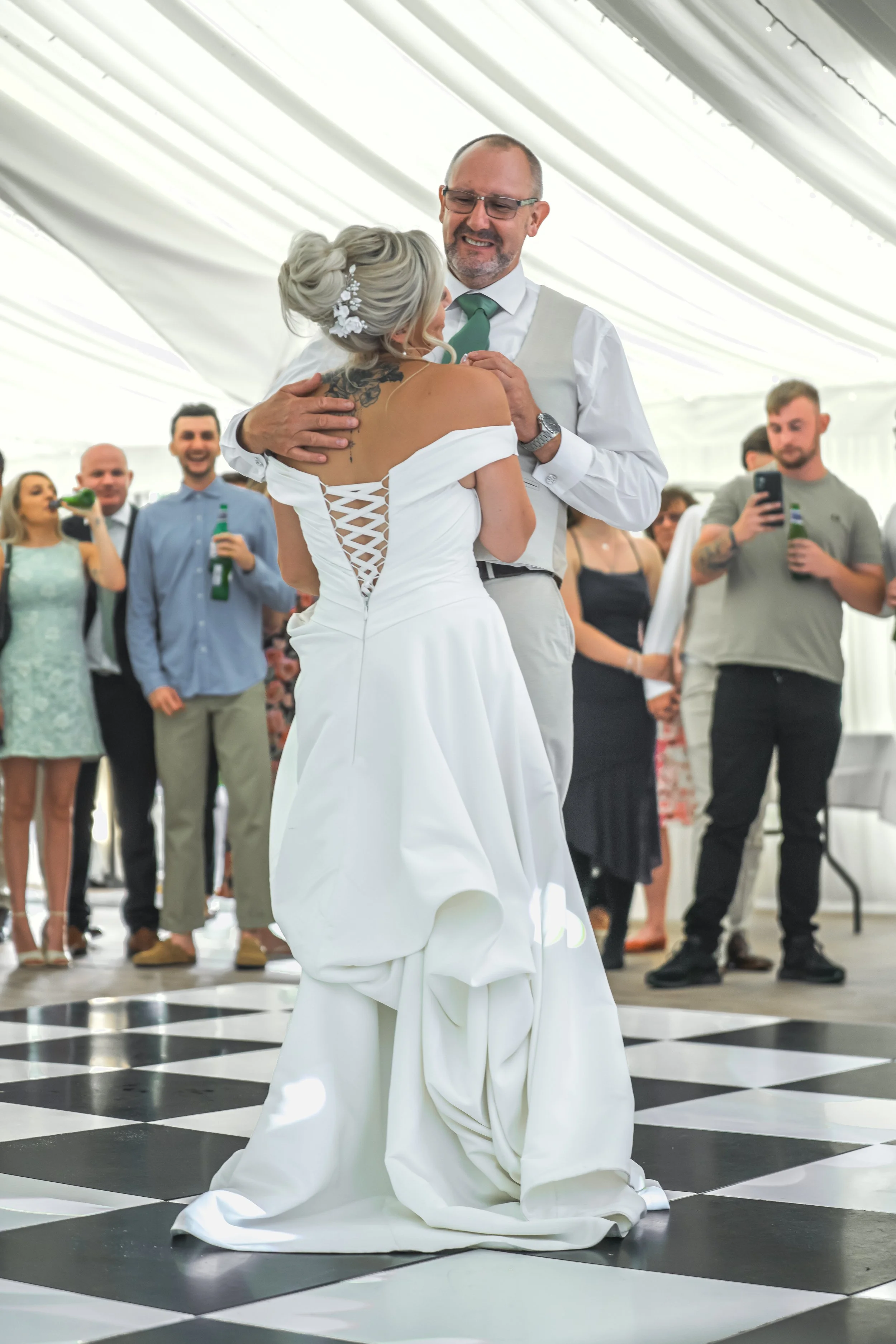 A bride and groom are dancing at a wedding reception under a white tent, with guests standing around and taking photos.