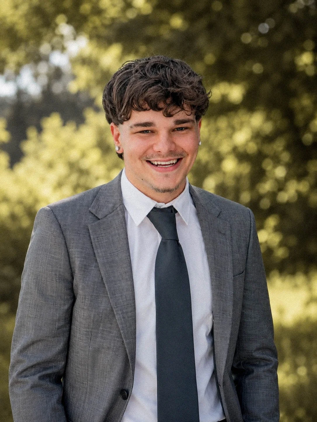 Young man with curly hair smiling outdoors, wearing a gray suit, white shirt, and black tie.
