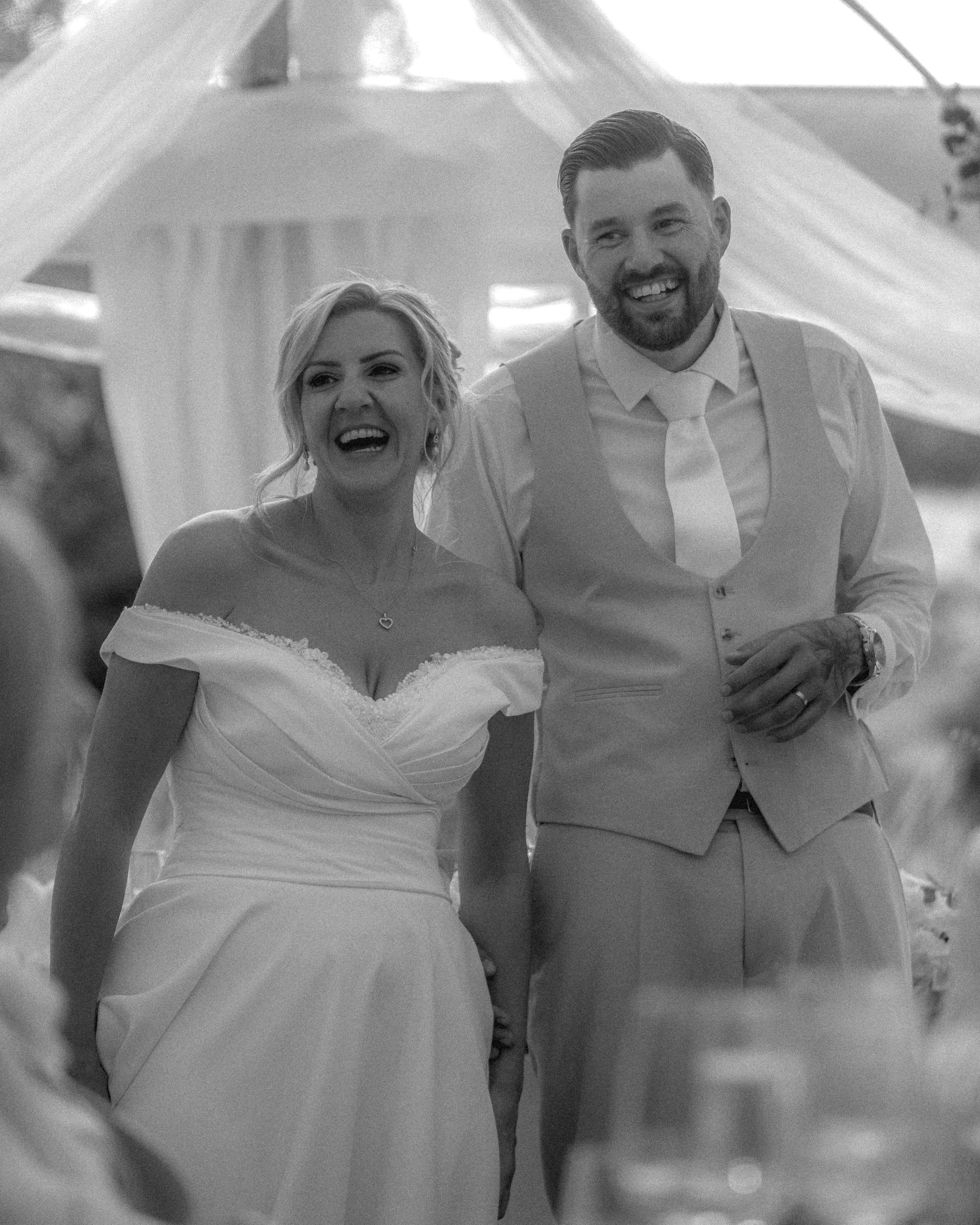 A man and woman dressed in wedding attire smiling and looking at each other. The woman is in a wedding dress and the man in a suit with suspenders and a tie, in a festive setting, likely a wedding reception.