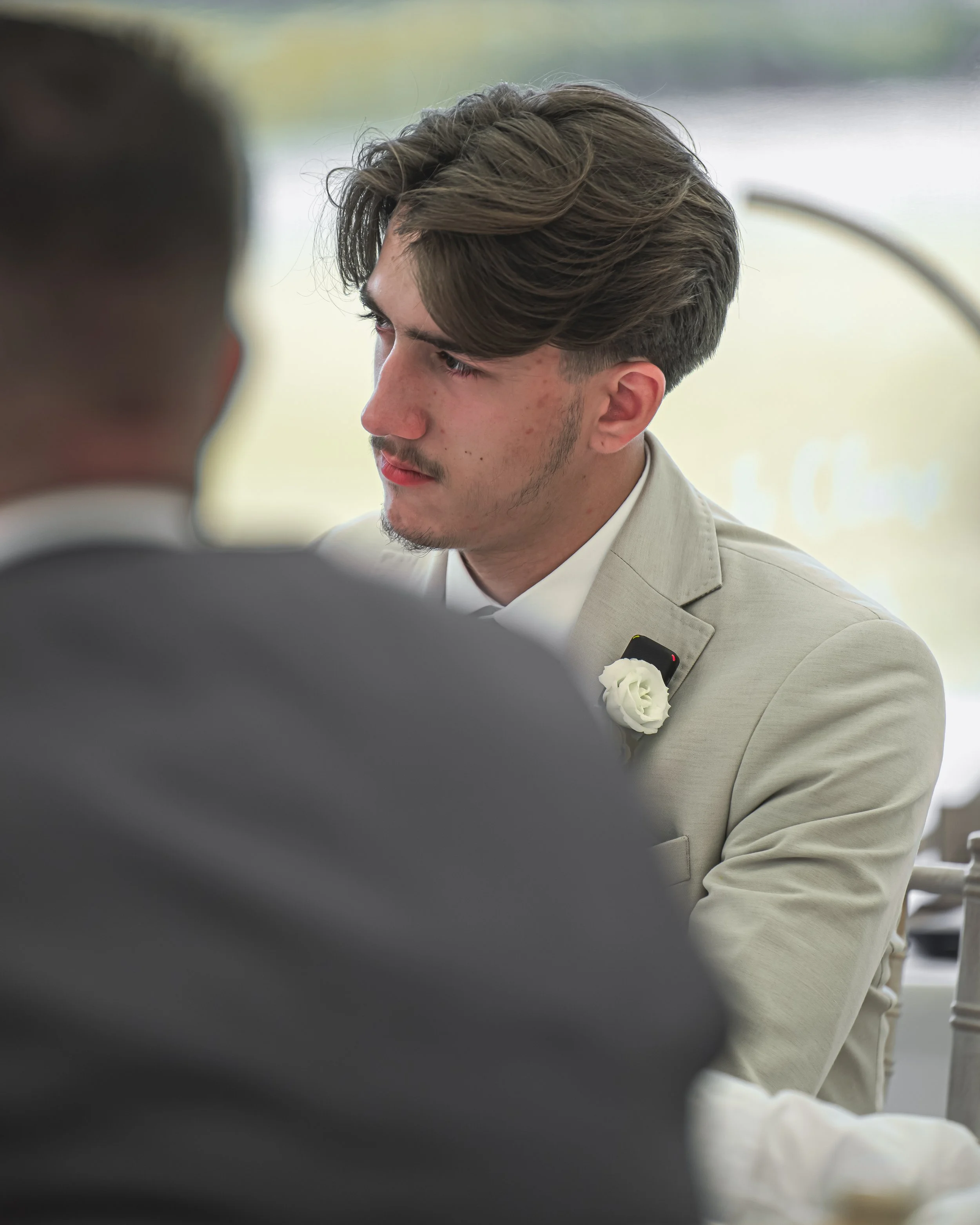 A close-up of a young man with brown hair styled to the side, dressed in a light-colored suit with a white shirt, sitting in a well-lit environment with a blurred background. The focus is on his face and upper body, with another person in the foregro