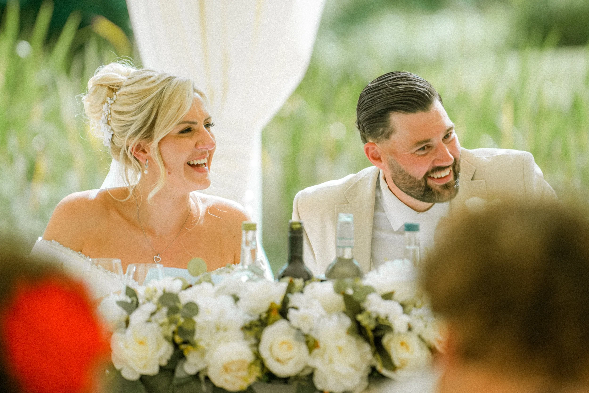 A bride and groom smiling during their wedding reception, with a table of white floral centerpieces and multiple bottles in front of them.