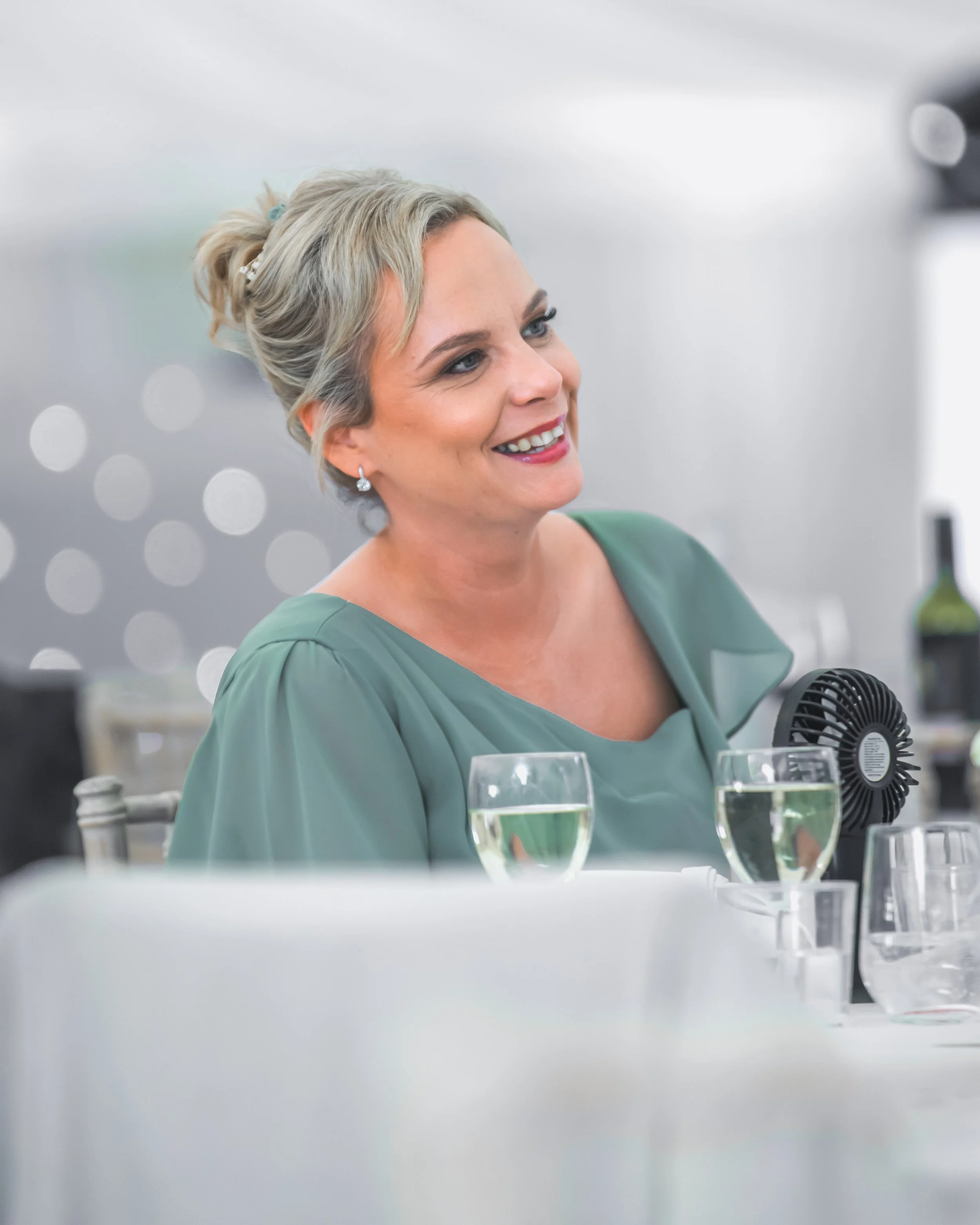 A woman with short blonde hair, earrings, and makeup, smiling while sitting at a table with glasses of champagne and a small black fan, in a well-lit indoor setting.