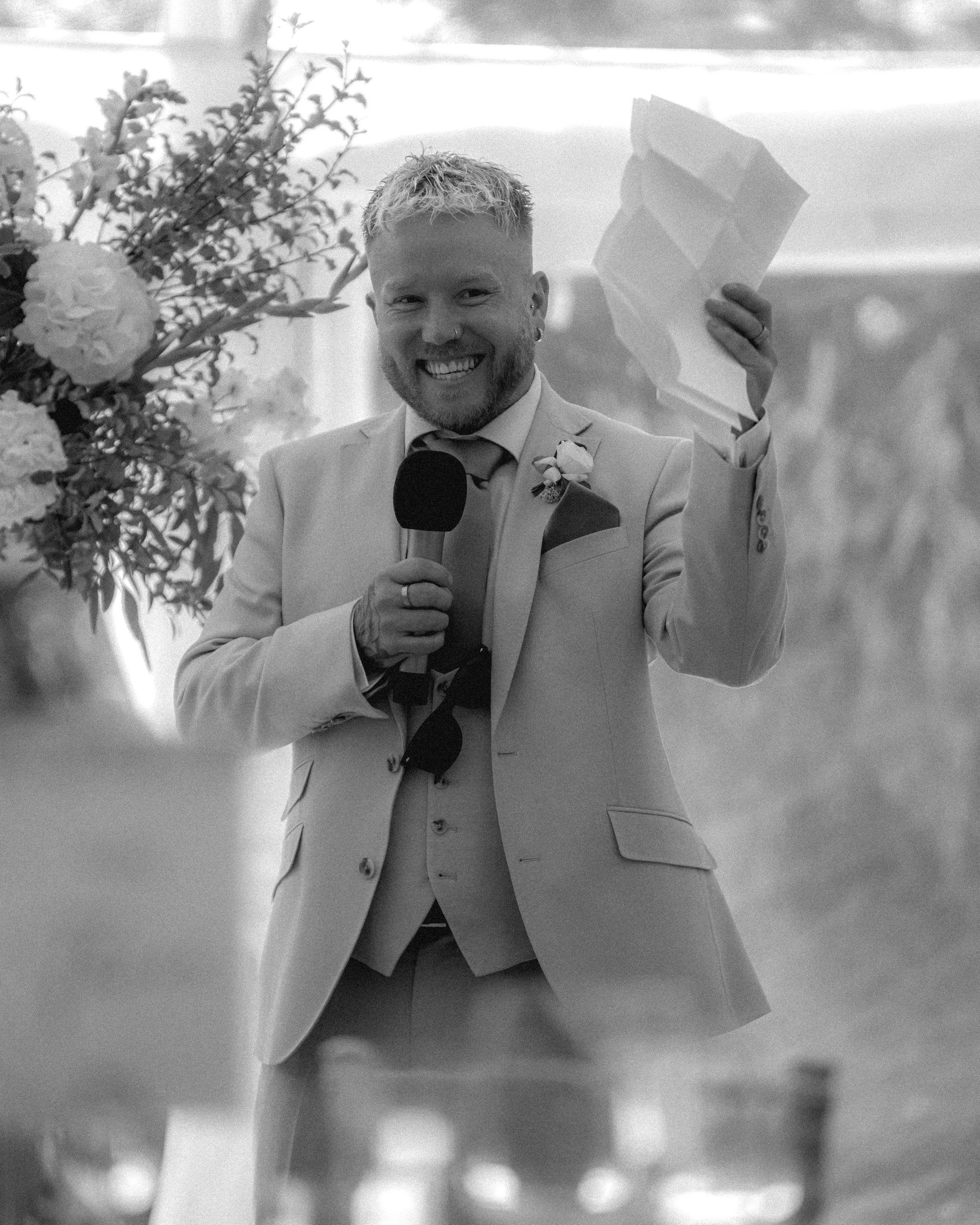 A man in a suit smiling and holding a microphone, standing behind a newspaper, with flowers on the side, indoor setting.