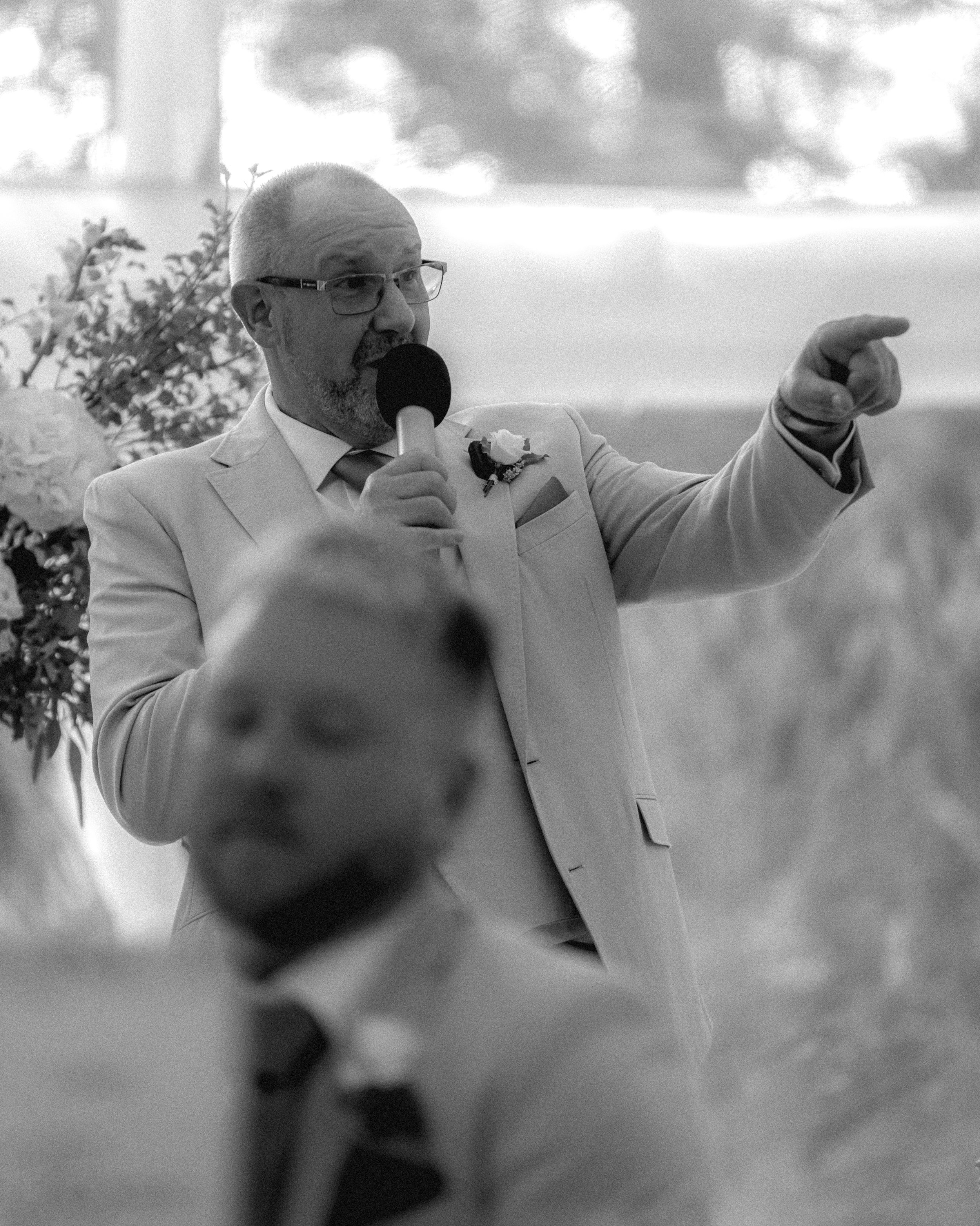 A man in a light-colored suit and glasses holding a microphone, pointing with his finger, with floral decorations in the background, at what appears to be an indoor event.