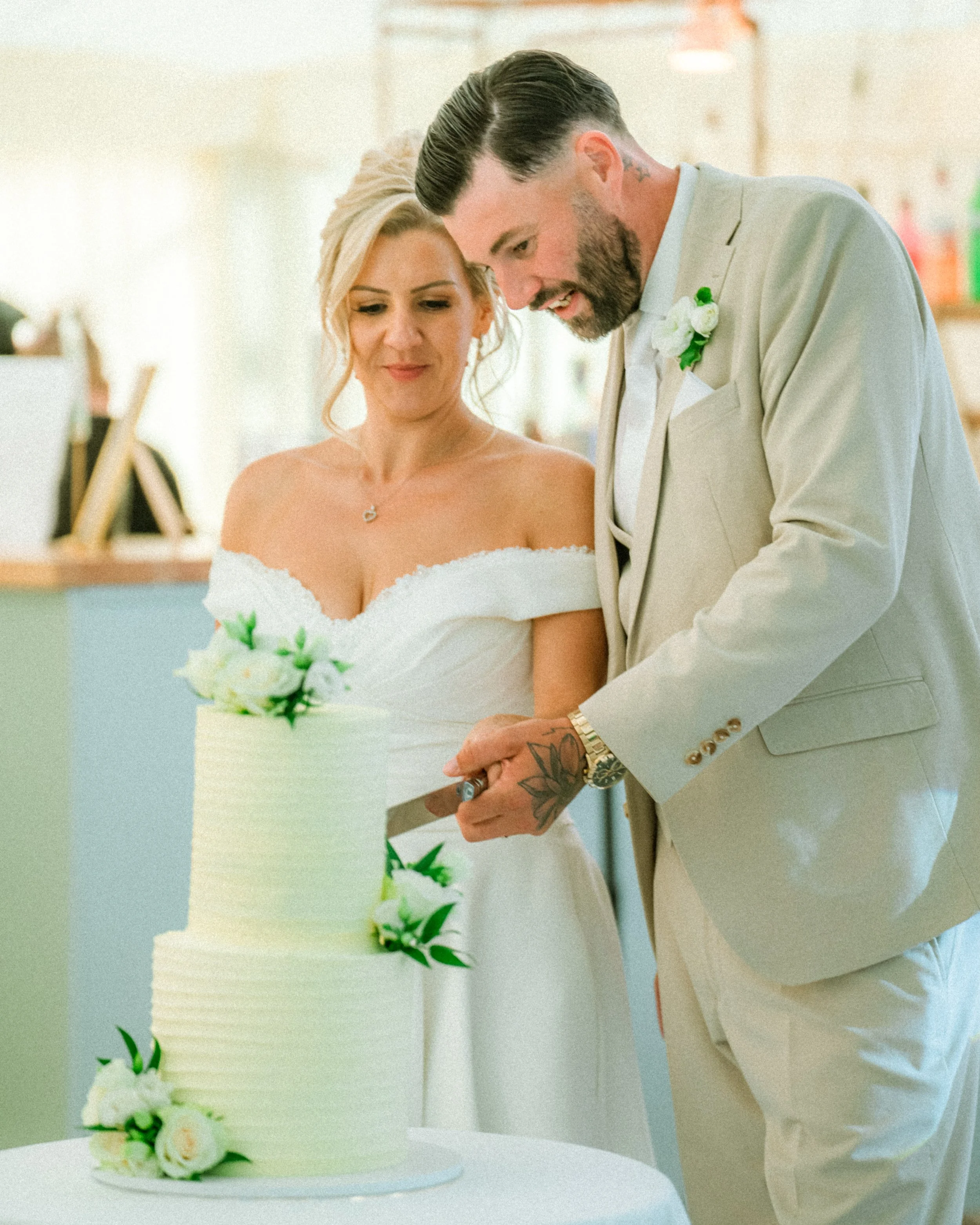 A bride and groom cutting their wedding cake together. The bride is wearing an off-the-shoulder wedding dress, and the groom is in a beige suit with a boutonnière. They are looking at the cake, which is decorated with white and green flowers, as they cut it.
