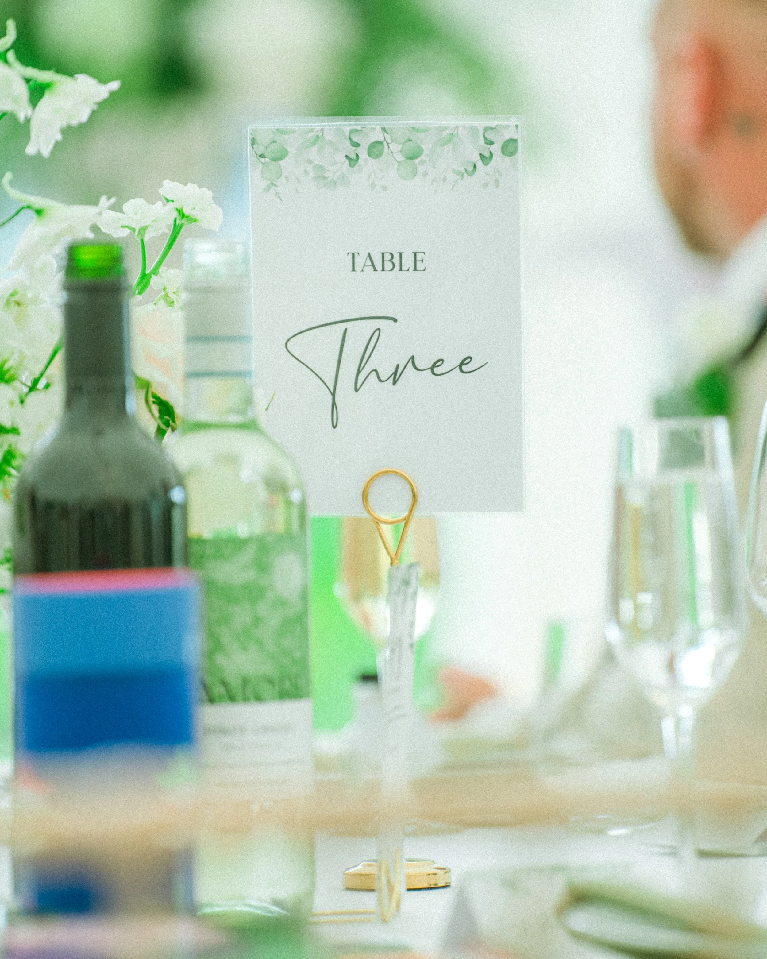 A table marker sign that reads 'Table Three' with a decorative leaf border at the top, set among bottles and flowers in the background.