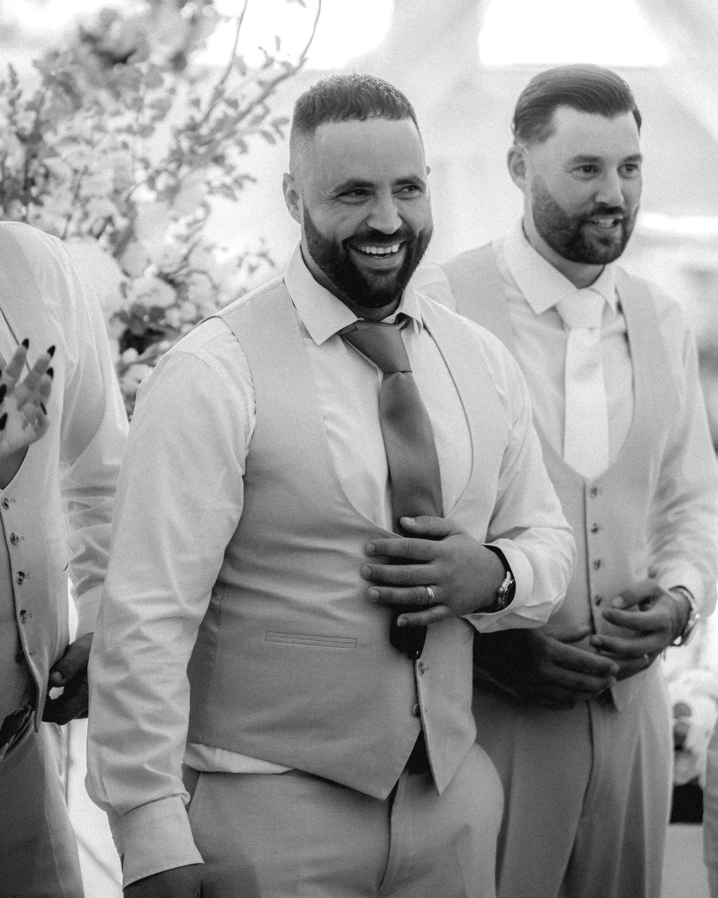 Two men in formal attire are standing side by side in a setting with other people. The man on the left has a beard, short hair, and is smiling widely. He is wearing a white shirt, a tie, and a vest. The man on the right has a beard and slicked-back h