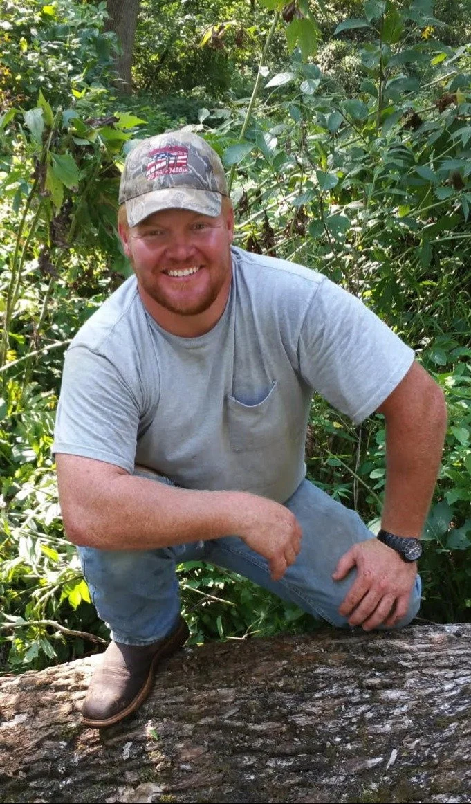 A smiling man kneeling on a fallen tree in a dense, green forest, wearing a camouflage baseball cap, gray t-shirt, jeans, and a watch, surrounded by lush foliage.