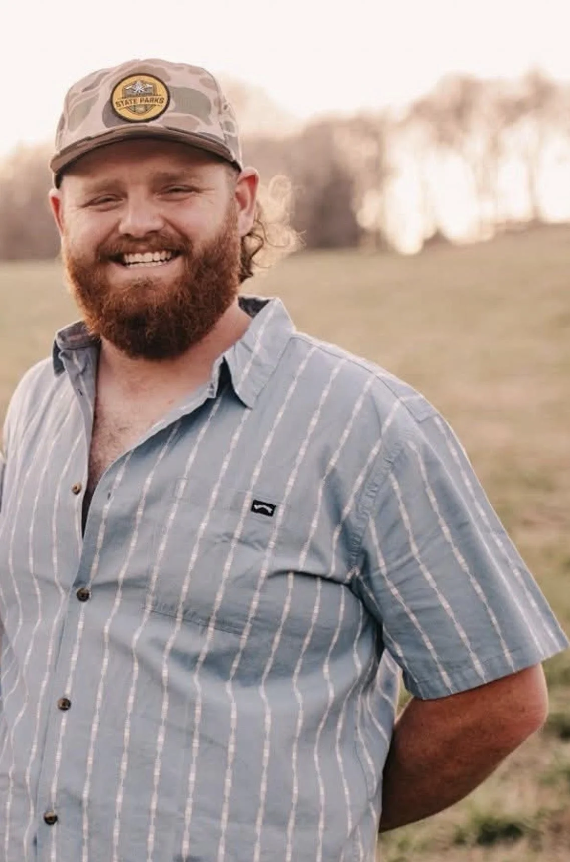 A man with a red beard wearing a gray baseball cap with a logo and a striped button-up shirt standing outdoors in a field with trees in the background.