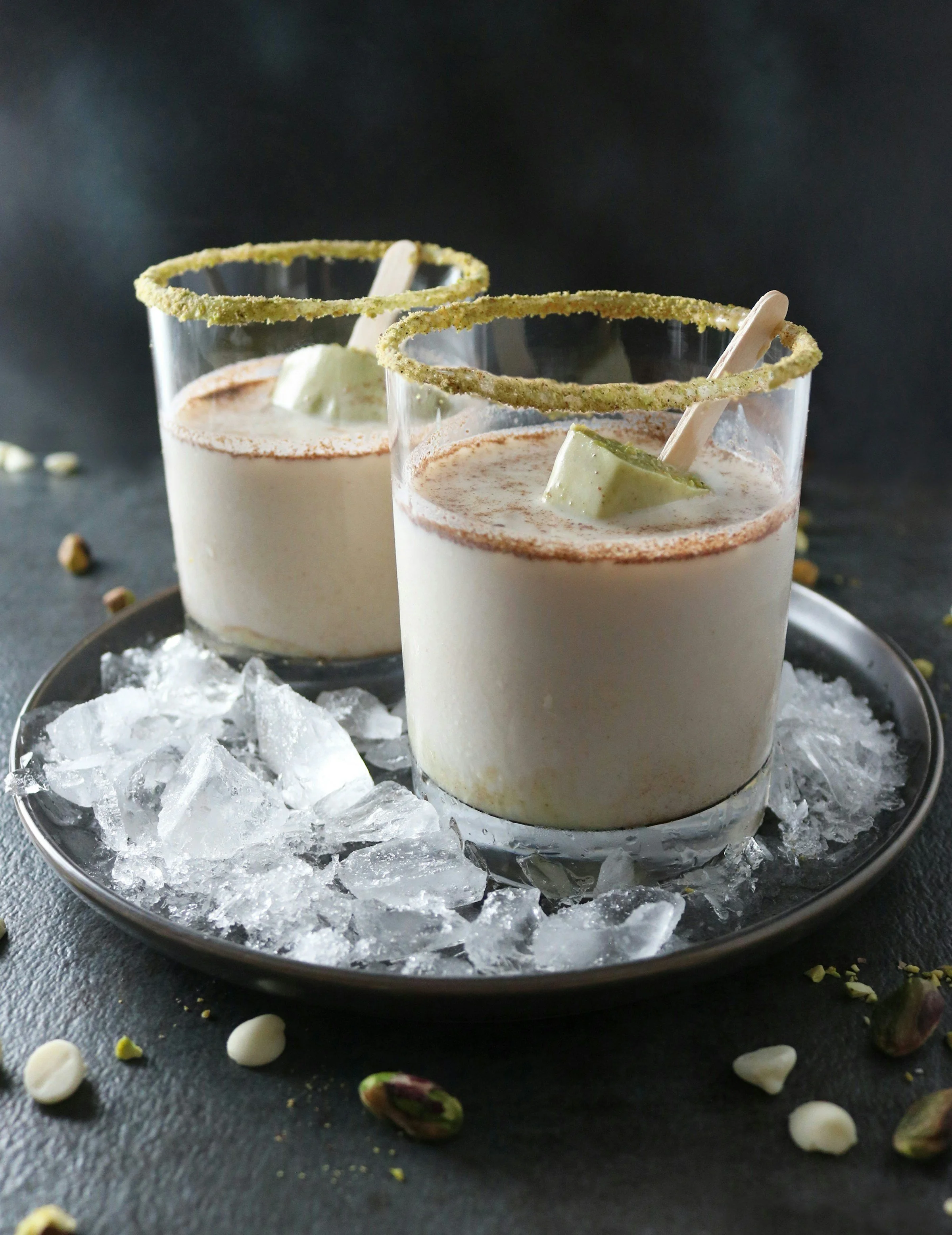 Two glasses of a creamy beverage garnished with lime wedges, with chocolate or coffee powder on top, surrounded by crushed ice in a metal dish, with crushed pistachios scattered on a dark surface.