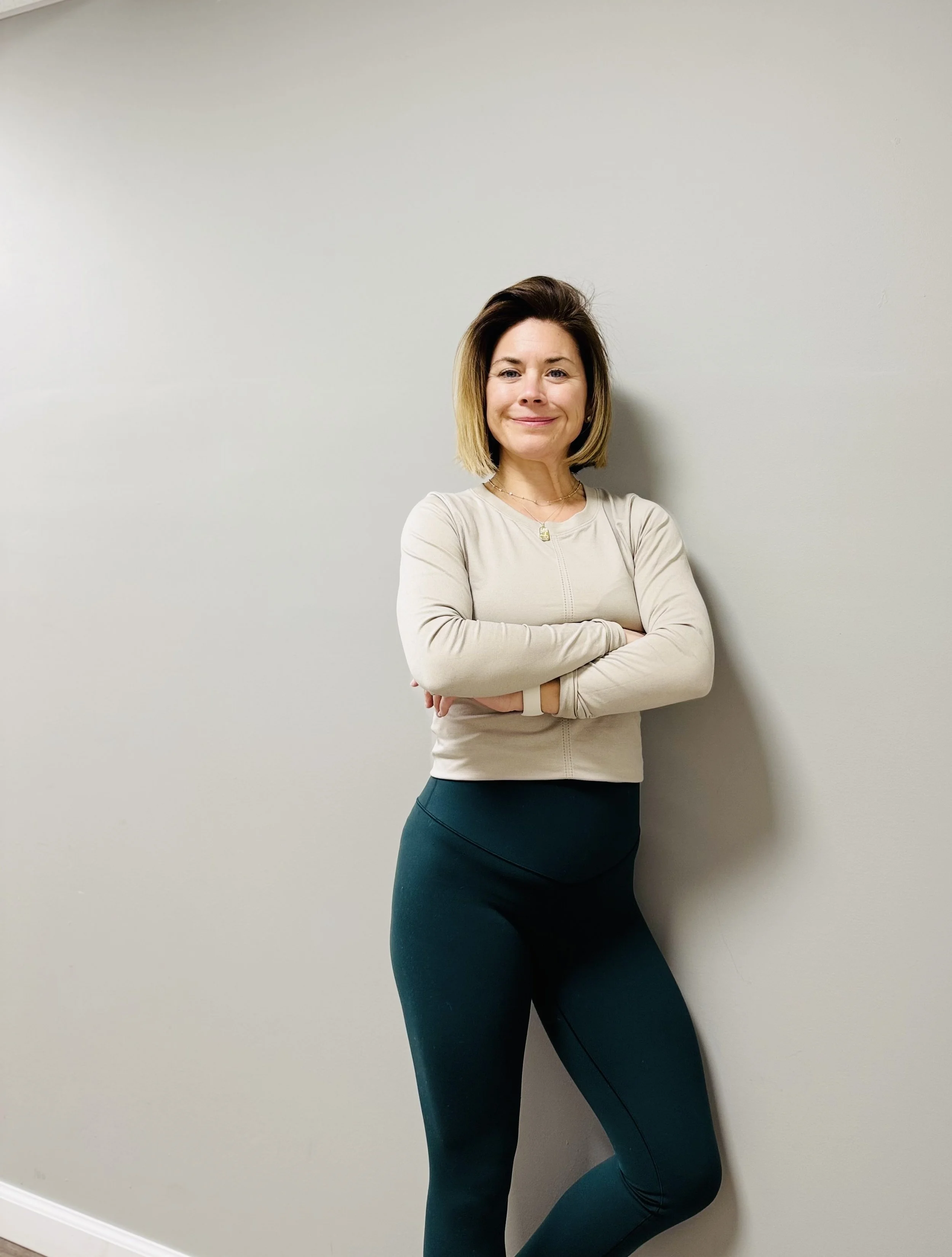 A woman with shoulder-length blonde hair, smiling confidently, standing against a plain light gray wall. She is crossing her arms and wearing a beige long-sleeve top and dark teal leggings.
