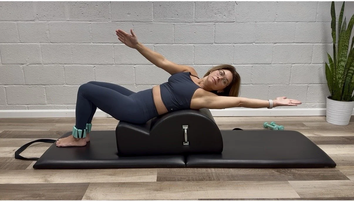Woman performing a stretch or exercise on a Pilates arc and a mat in a fitness studio with a potted plant in the background.