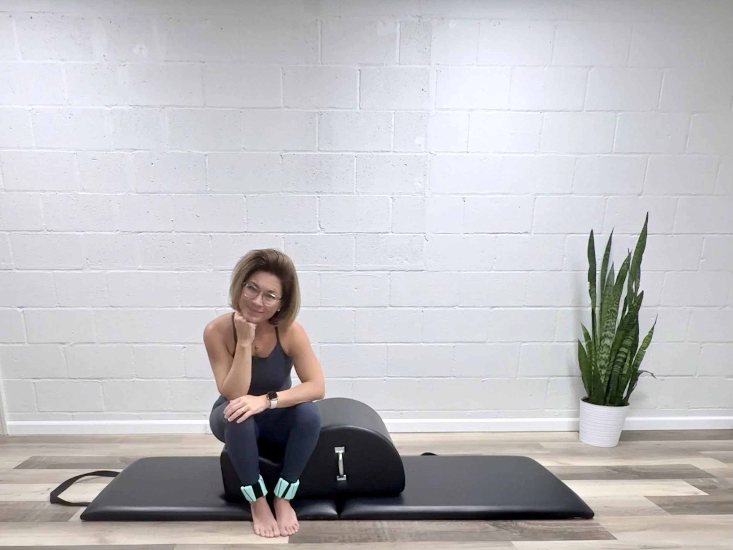 A woman with glasses, wearing a black tank top, gray pants, and light blue ankle weights, sitting on a Pilates spine corrector on a black Pilates mat in a room with a white brick wall and a tall green plant in a white pot to her right.