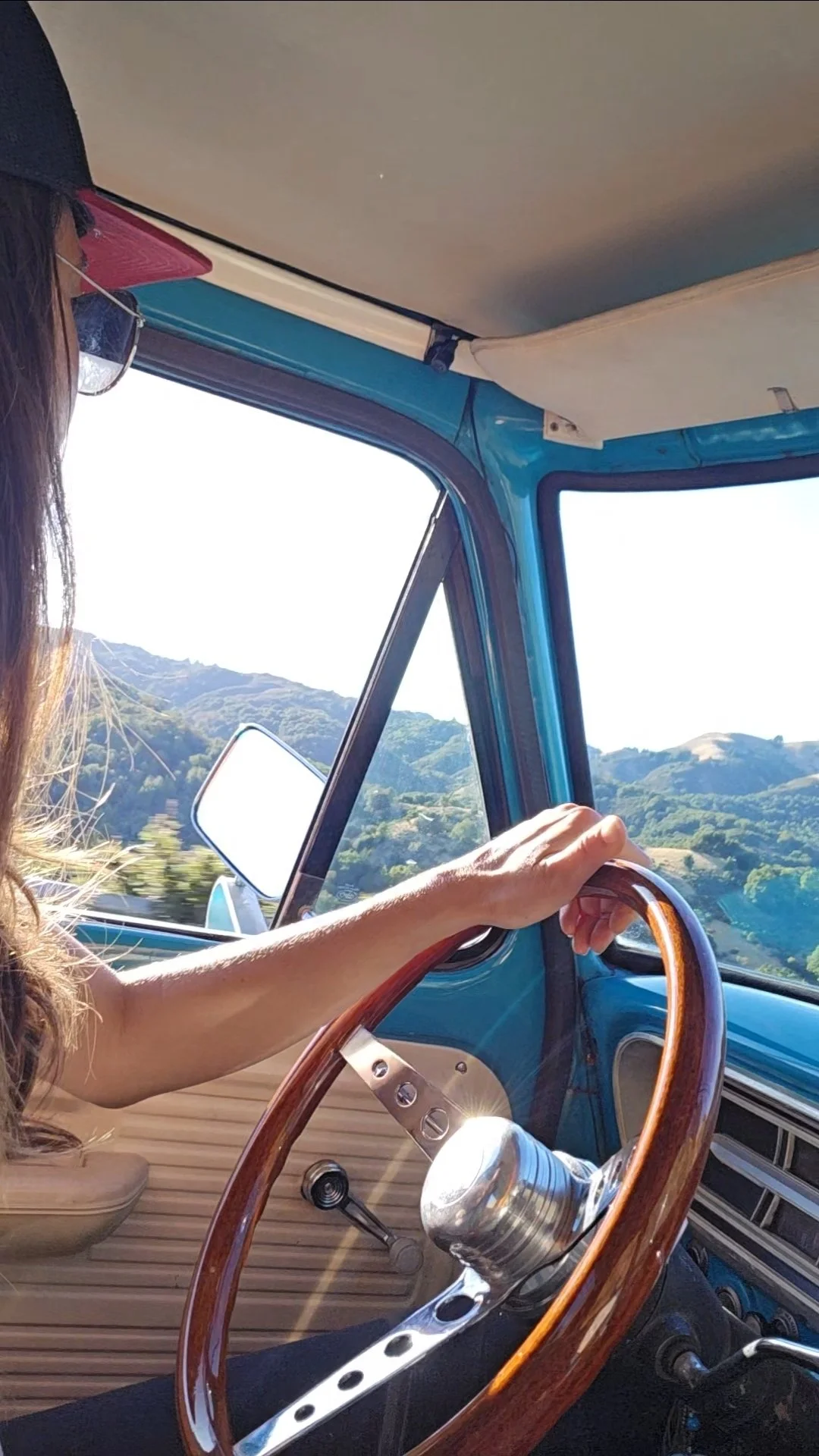 Person driving a vintage blue vehicle, with wooden steering wheel, in a scenic mountainous landscape.