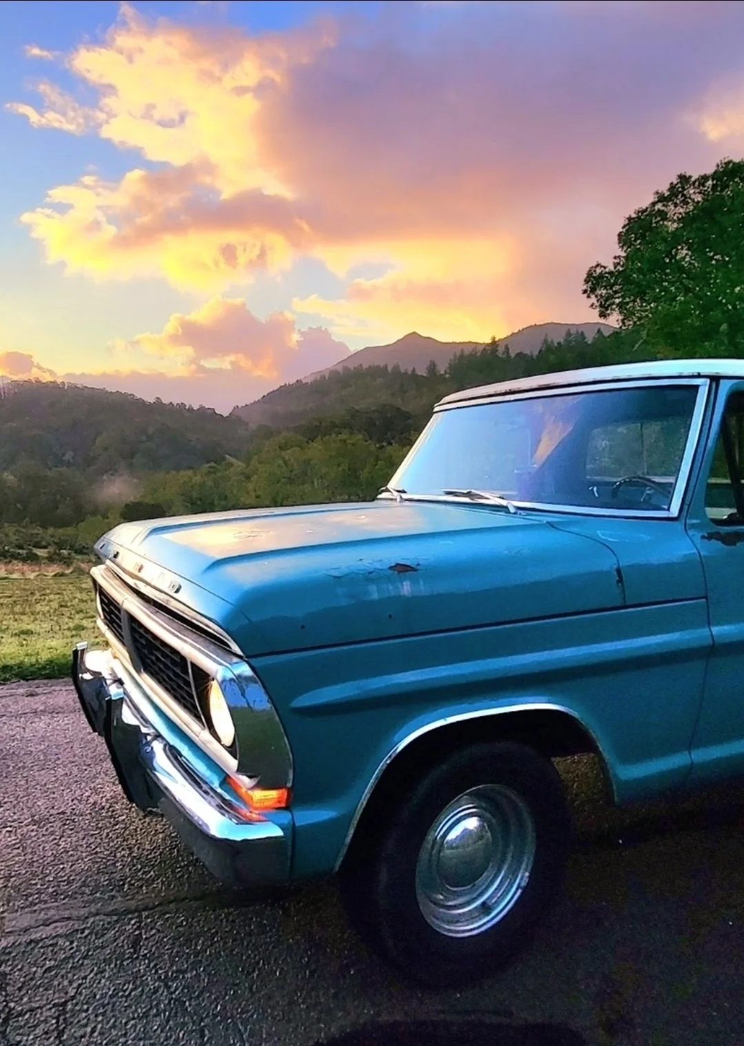 A vintage blue pickup truck parked on a road with a scenic mountain landscape and vibrant sunset sky in the background.