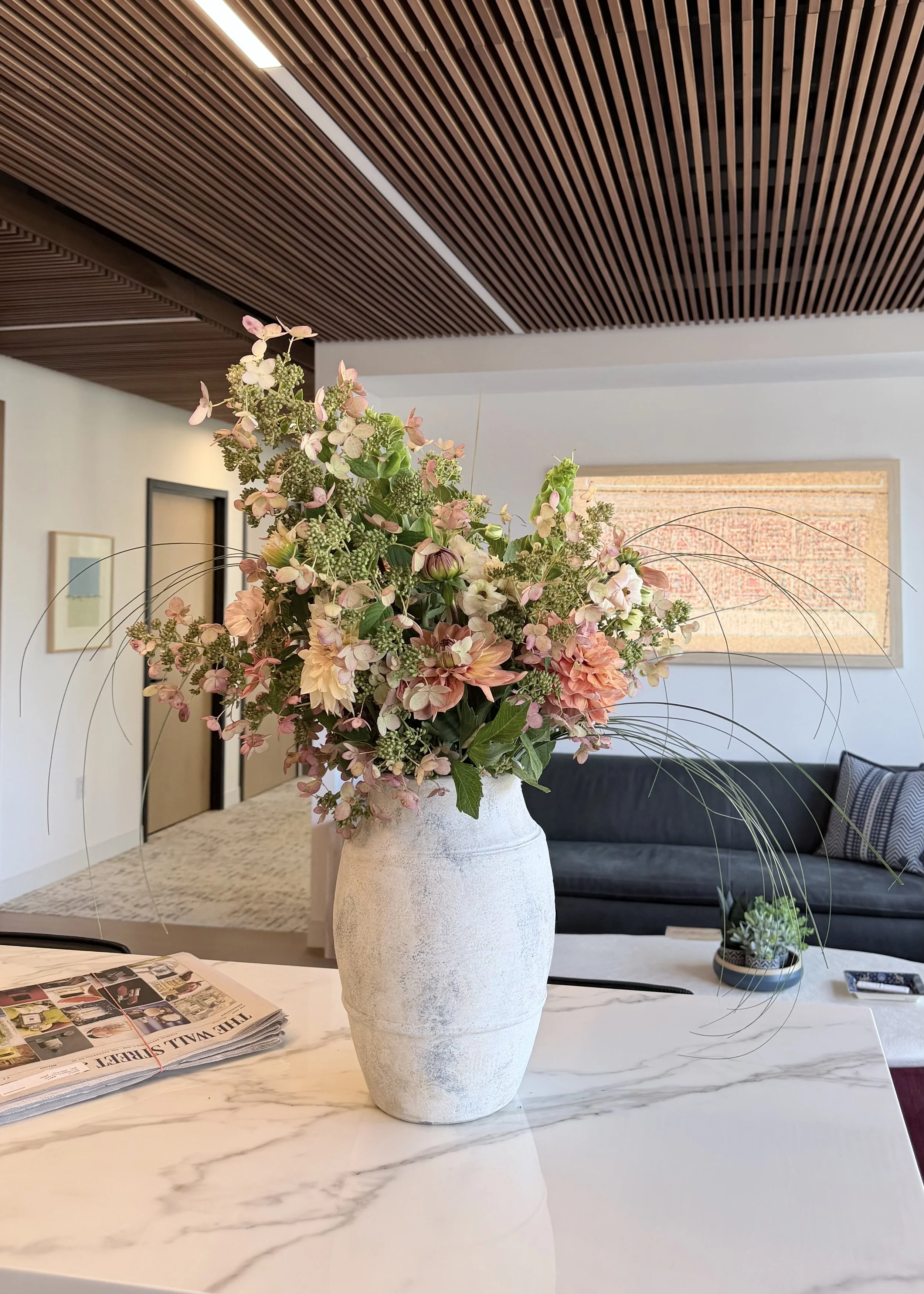 A large white vase with a textured surface holding a bouquet of pink, peach, and green flowers on a white marble table with gray veining. In the background, a black sofa with striped pillows, framed artwork on the wall, and a large textured artwork in a wooden frame are visible. A small potted plant is on the table, and a stack of newspapers is nearby.