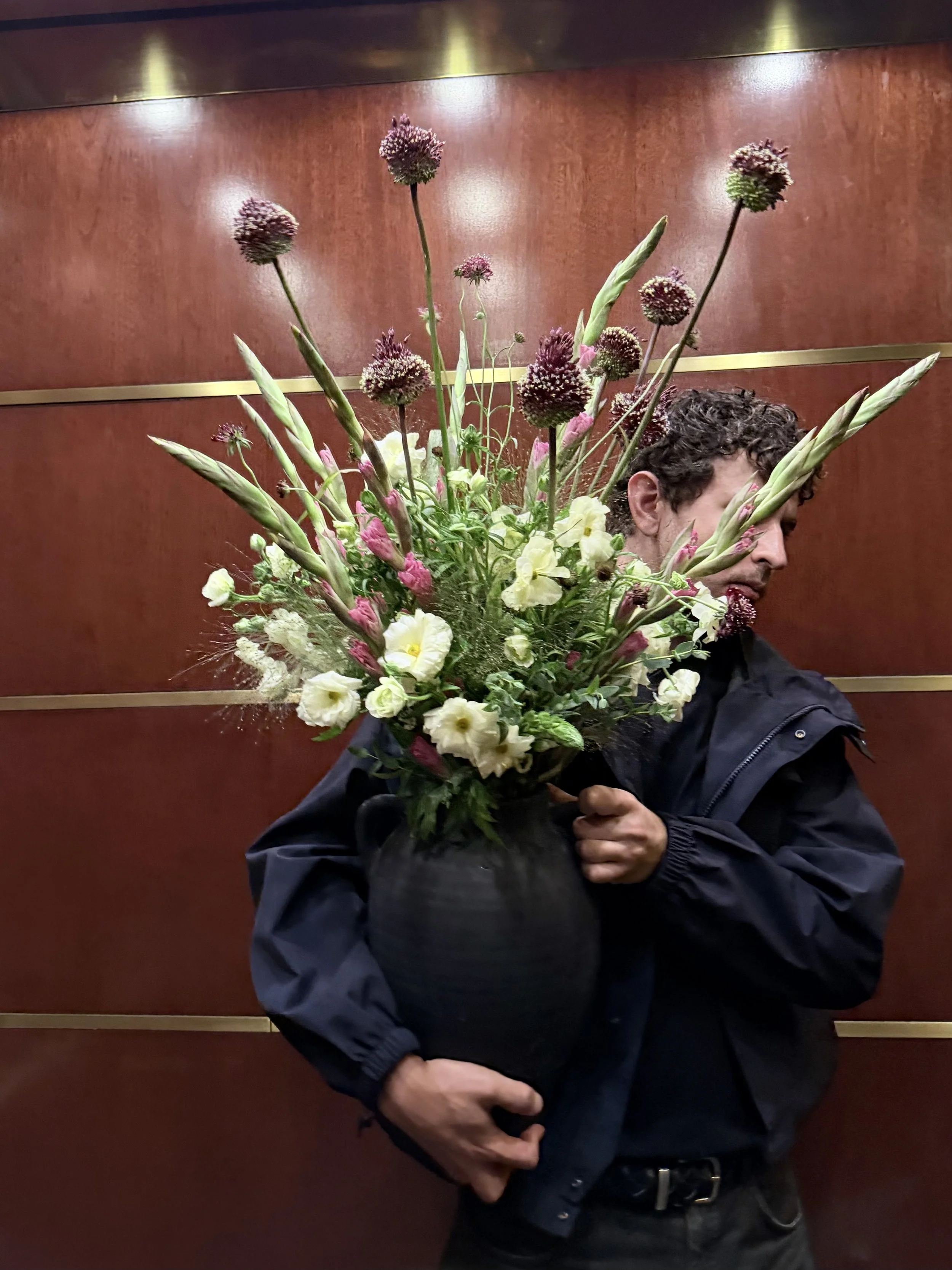 A man with curly dark hair holding a large bouquet of flowers in front of an elevator wall.