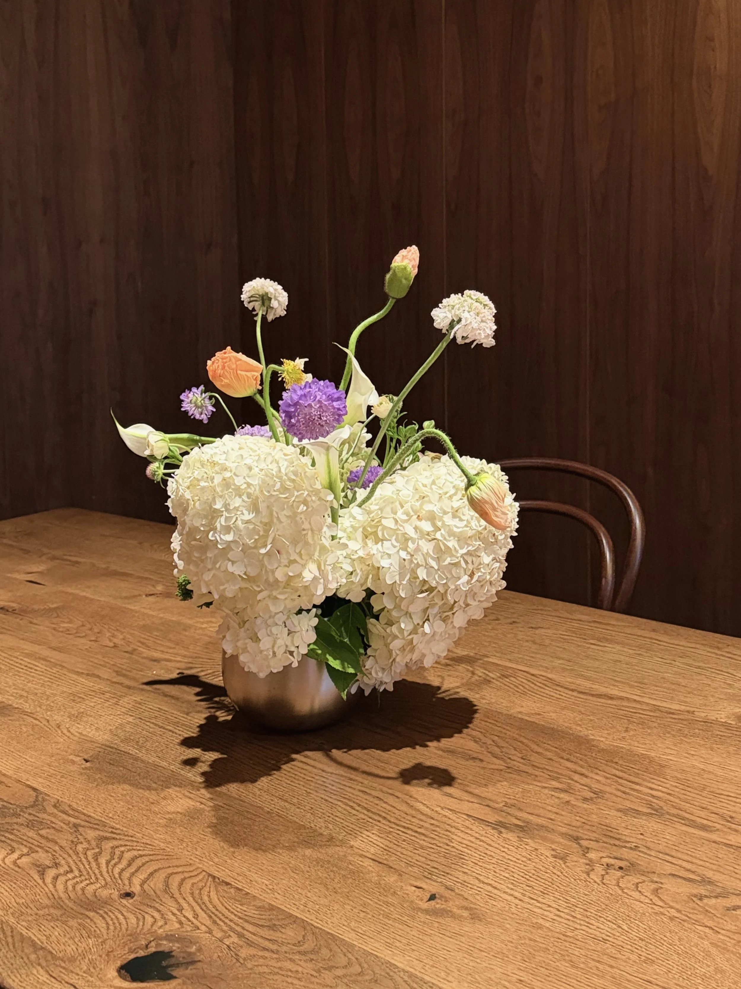 A floral arrangement with white hydrangeas, purple, orange, pink, and white flowers in a metallic vase, placed on a wooden table with a dark wooden background.