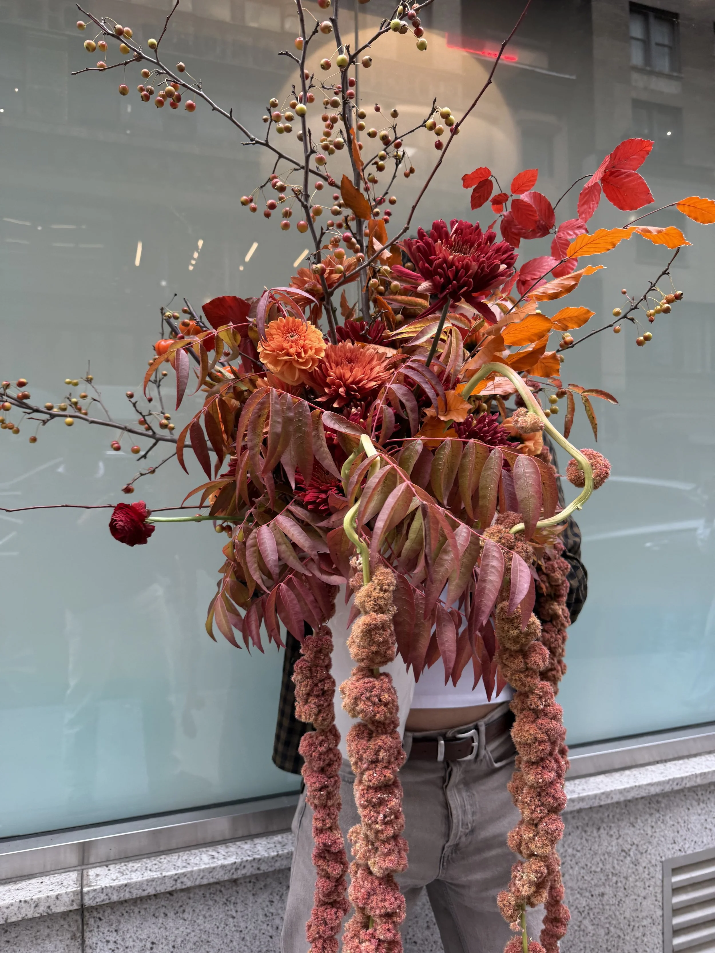 A person holding a large colorful flower arrangement in front of their body, with their head not visible. The arrangement includes branches with small berries, red and orange flowers, and long hanging fuzzy pink and orange pom-pom like flowers. The person is wearing gray pants and a checkered shirt, standing against a modern building with glass windows.