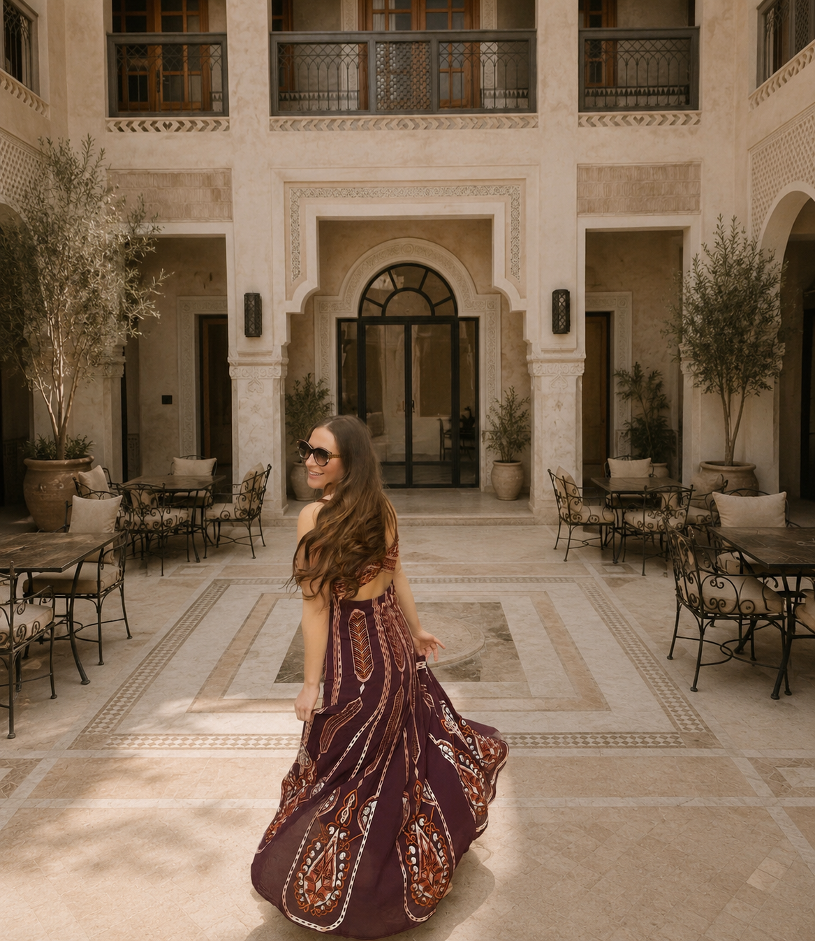 Woman in a patterned dress and sunglasses smiling in a courtyard with stone walls, potted trees, and outdoor tables and chairs.