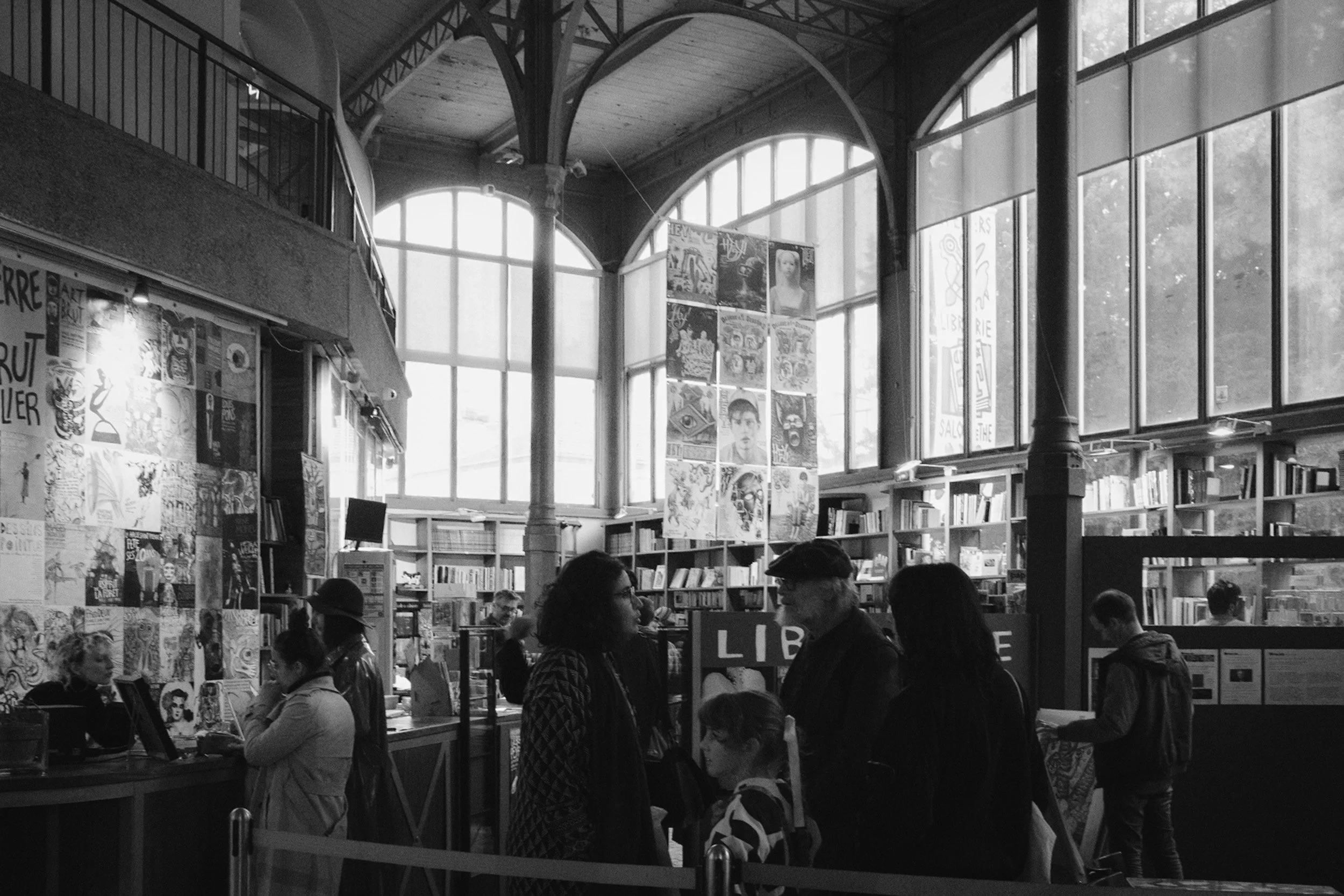A busy indoor bookstore or library with large stained glass windows, bookshelves, and posters on the wall, with people browsing and reading.