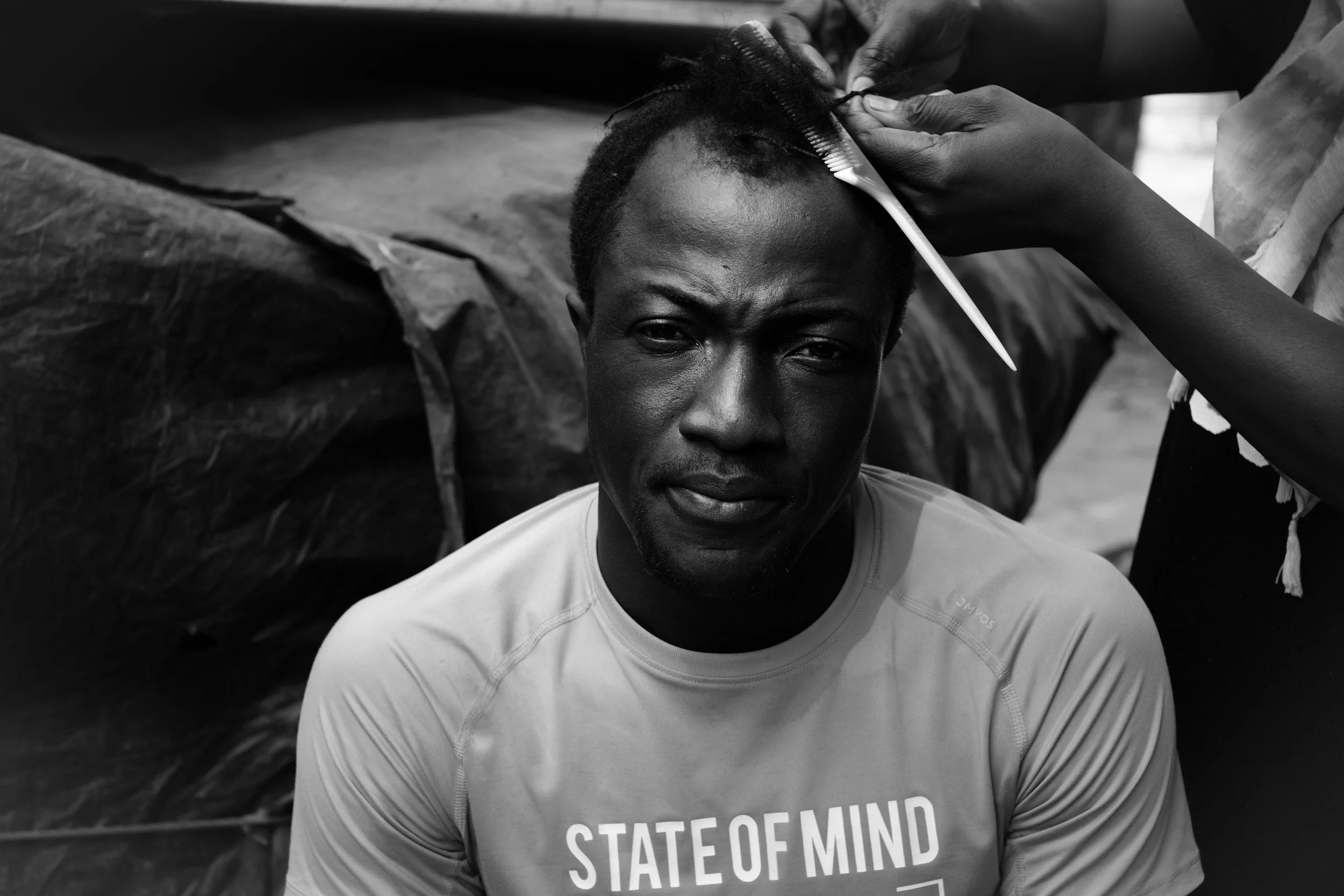 A person with short hair having their hairstyle cut by another individual. The person is wearing a T-shirt with the words 'STATE OF MIND' visible on it.