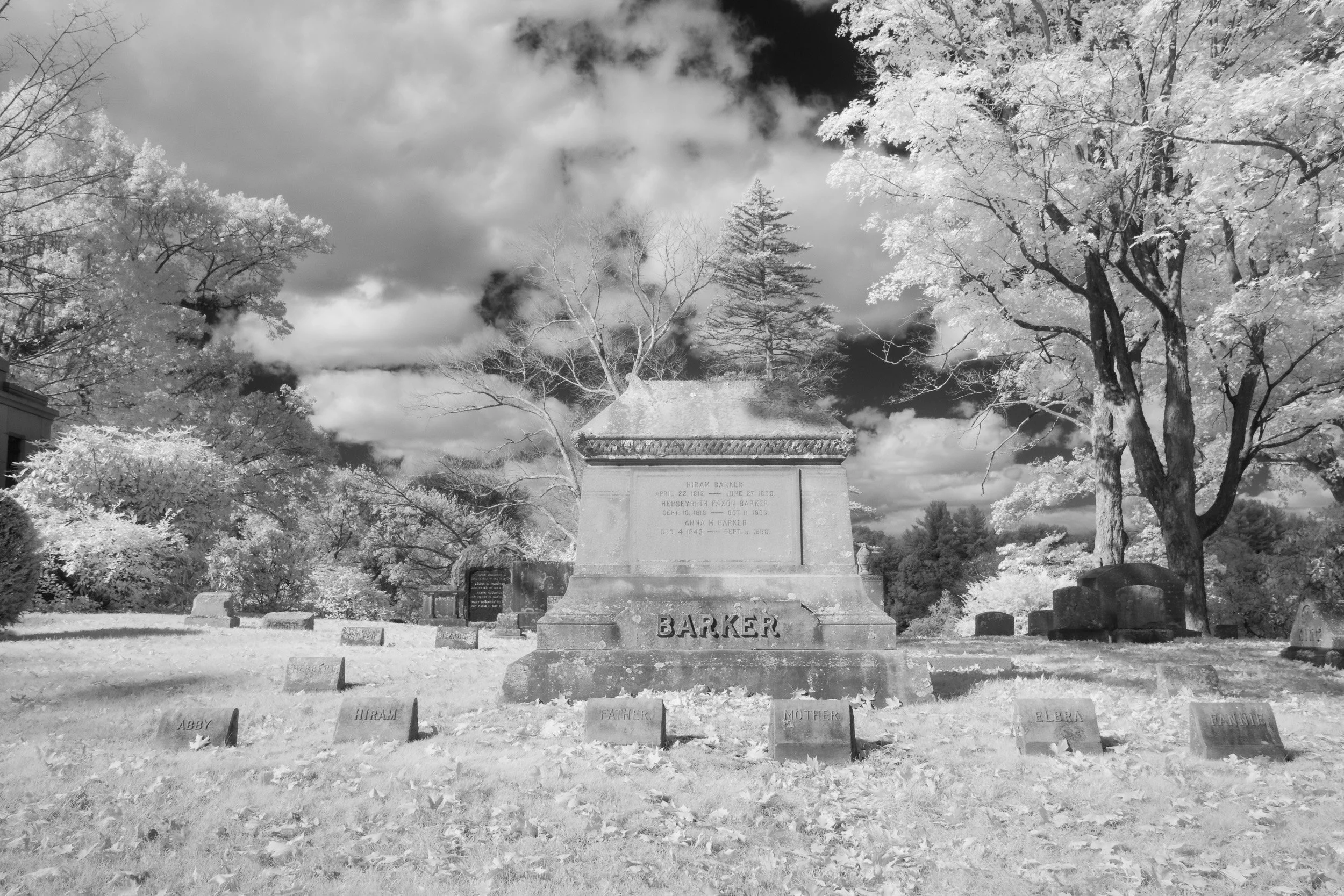 Gravestone in a cemetery with surrounding trees and a cloudy sky in the background. The gravestone has the name 'Barker' and a plaque mentioning 'Hiram Barker, April 22, 1812, June 27, 1895.'