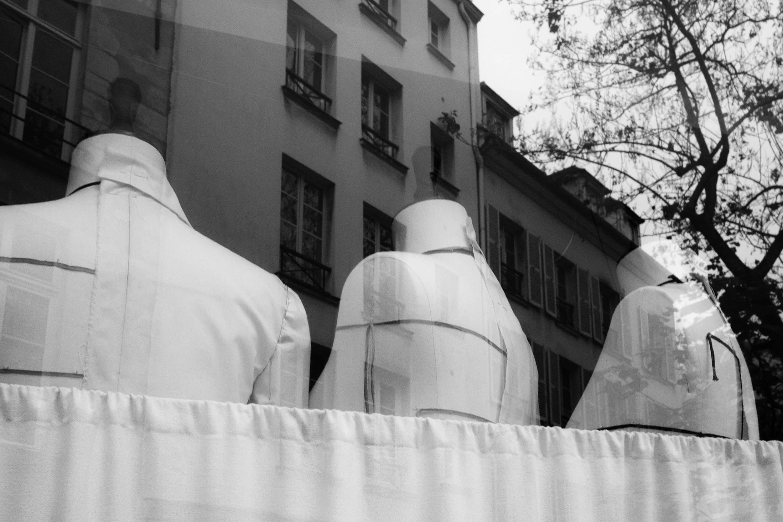 Black and white photo of city buildings with multiple windows, a tree with bare branches, and a cluster of white tents at the street level.