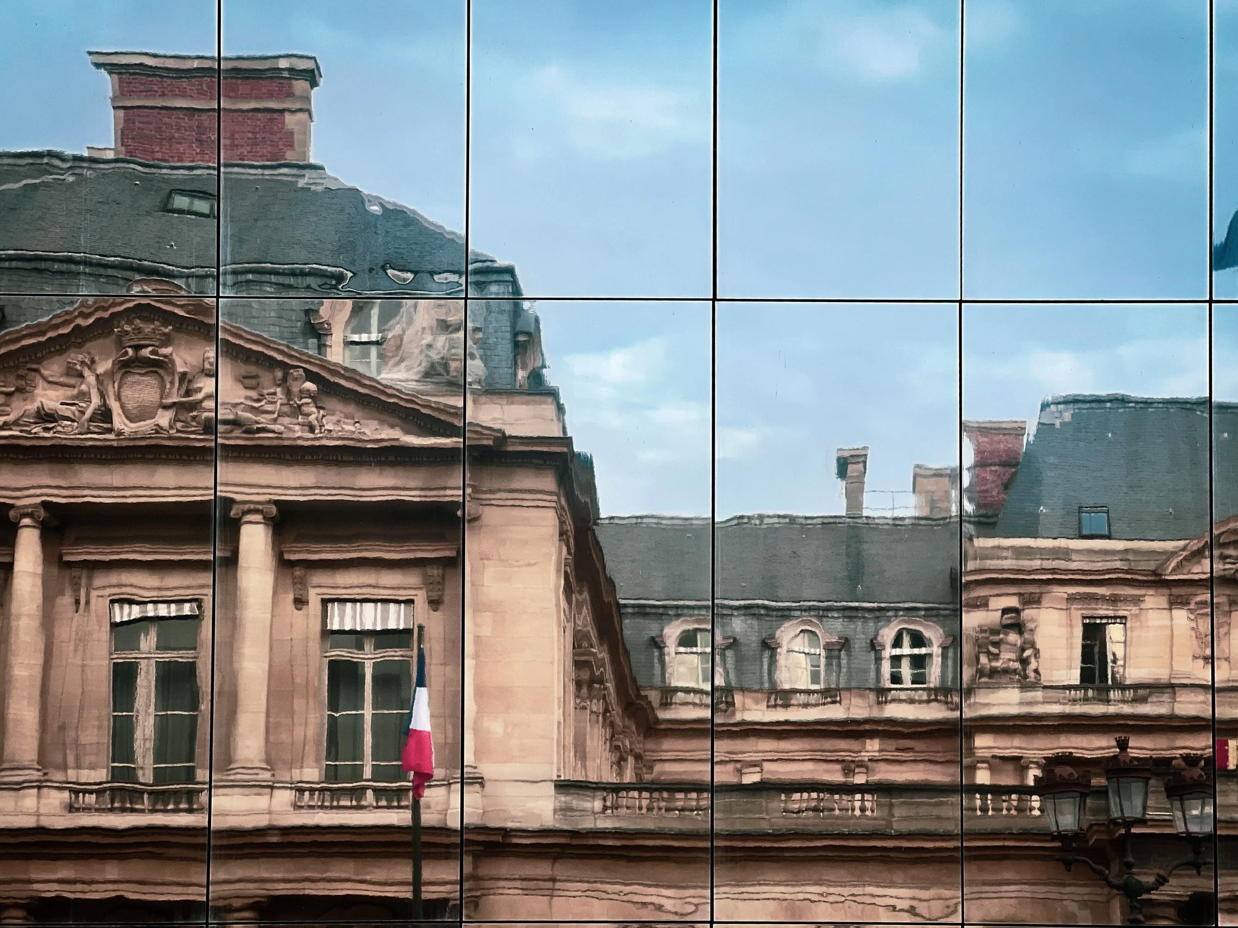 Reflection of an ornate historic building with classical architecture, columns, and decorative sculptures, seen on a glass surface with grid lines, under a cloudy sky.