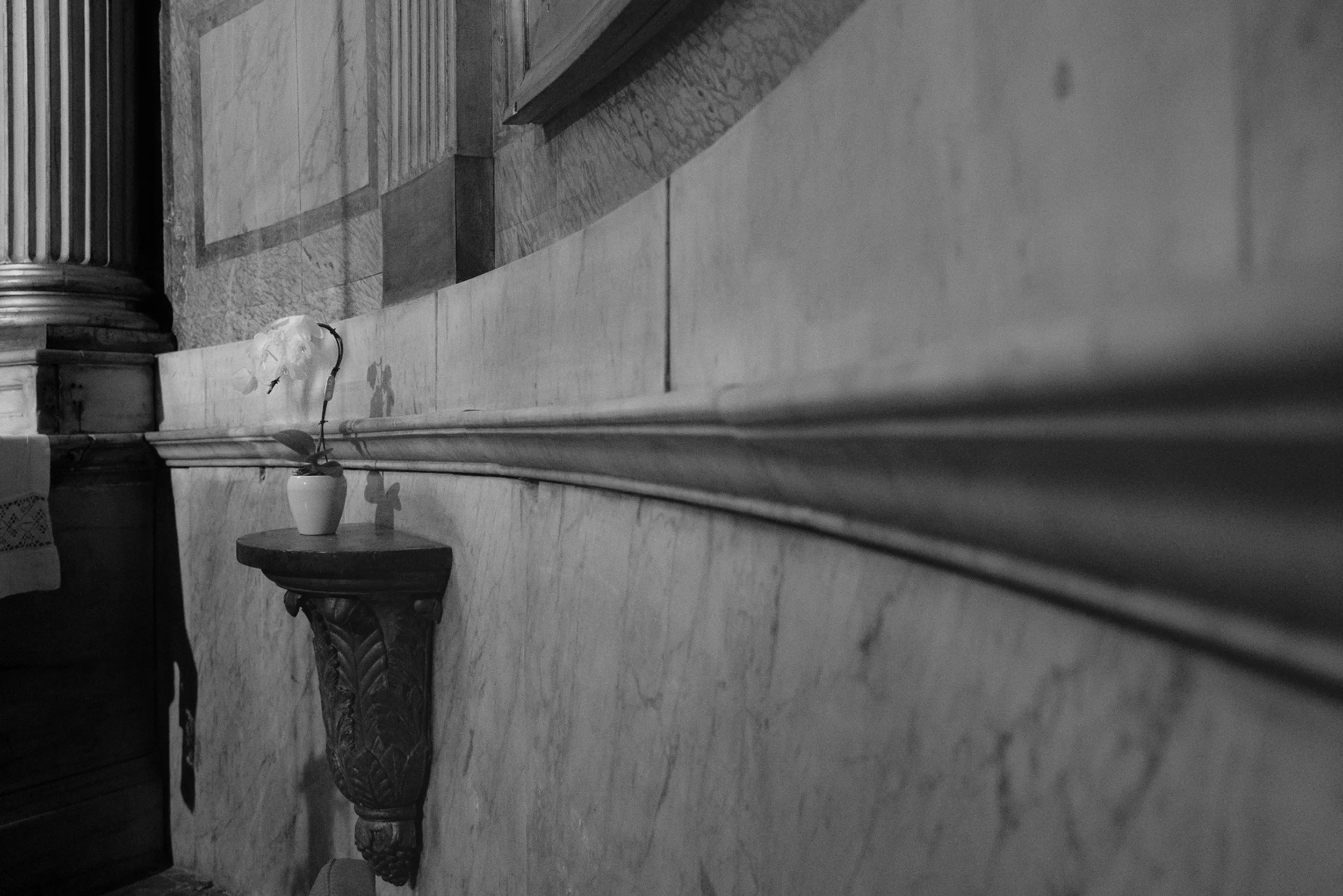 A hallway with a marble floor, a wooden staircase, a pot with a plant on a carved stand, and a column with fluted design.
